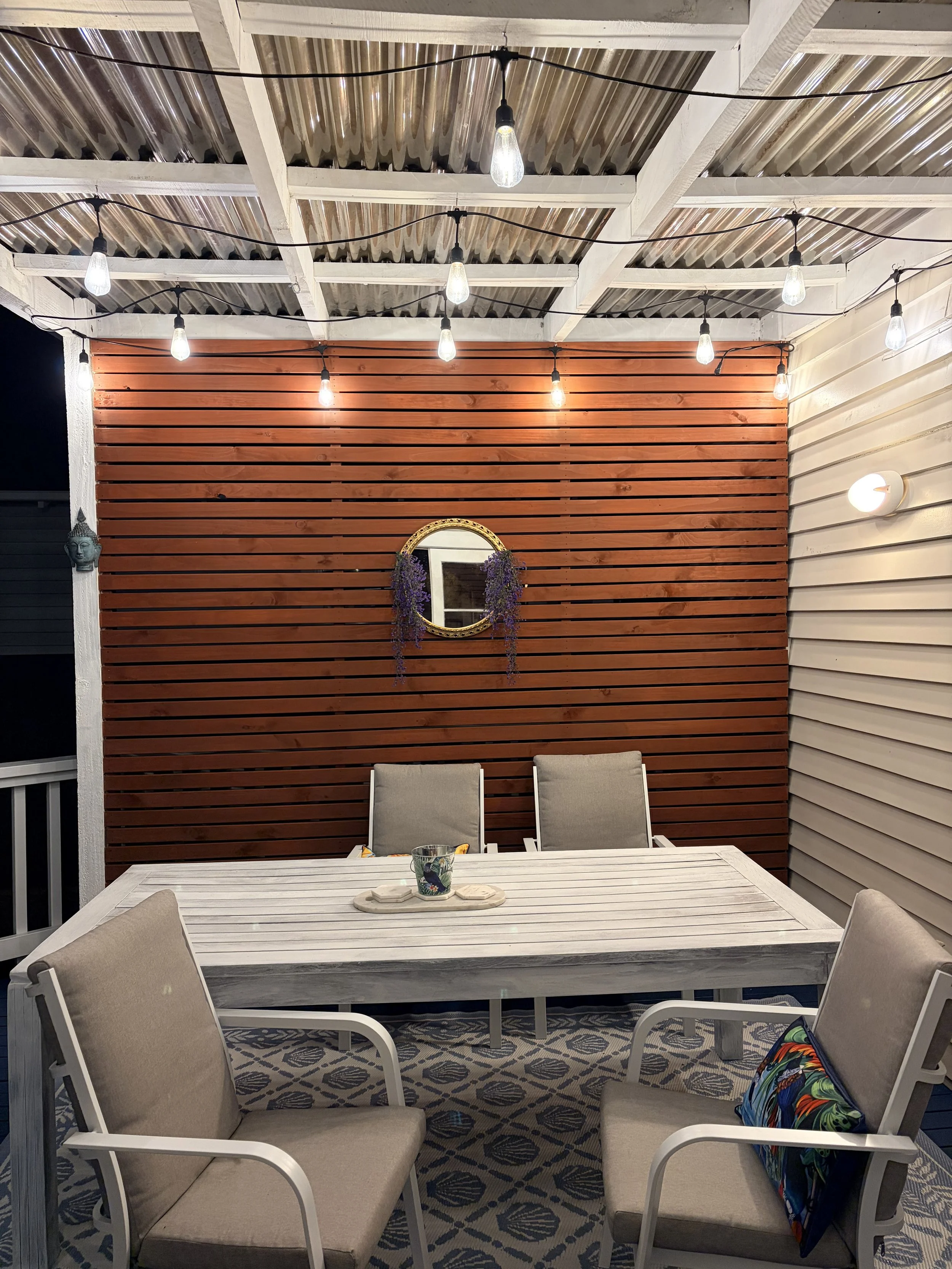 Outdoor patio area with a dining table, four beige chairs, string lights overhead, a wooden slat wall with a round mirror and purple flowers, a patterned rug, and a colorful bag on one chair.