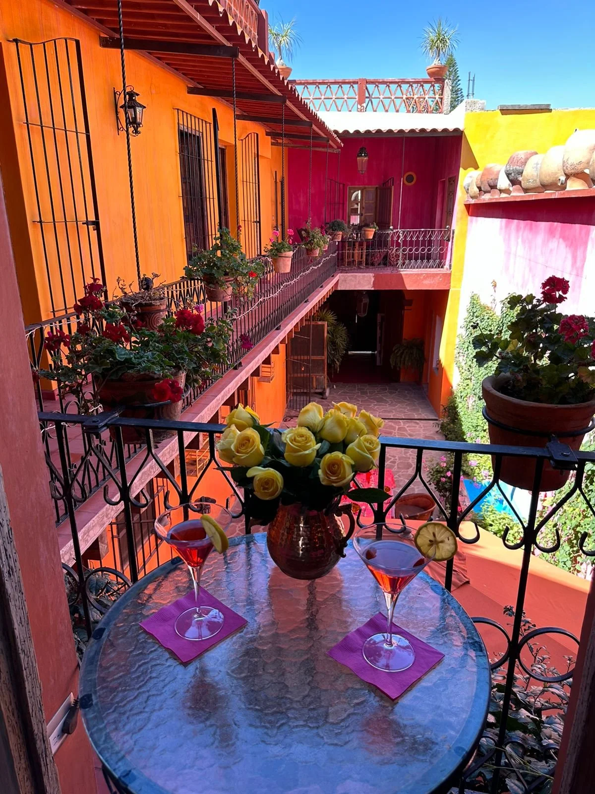 Vista de un balcón decorado con flores, con un mesa de cristal y dos copas con bebida y rodajas de limón, en un entorno colorido con paredes naranja, rosa y amarilla, y plantas en macetas.