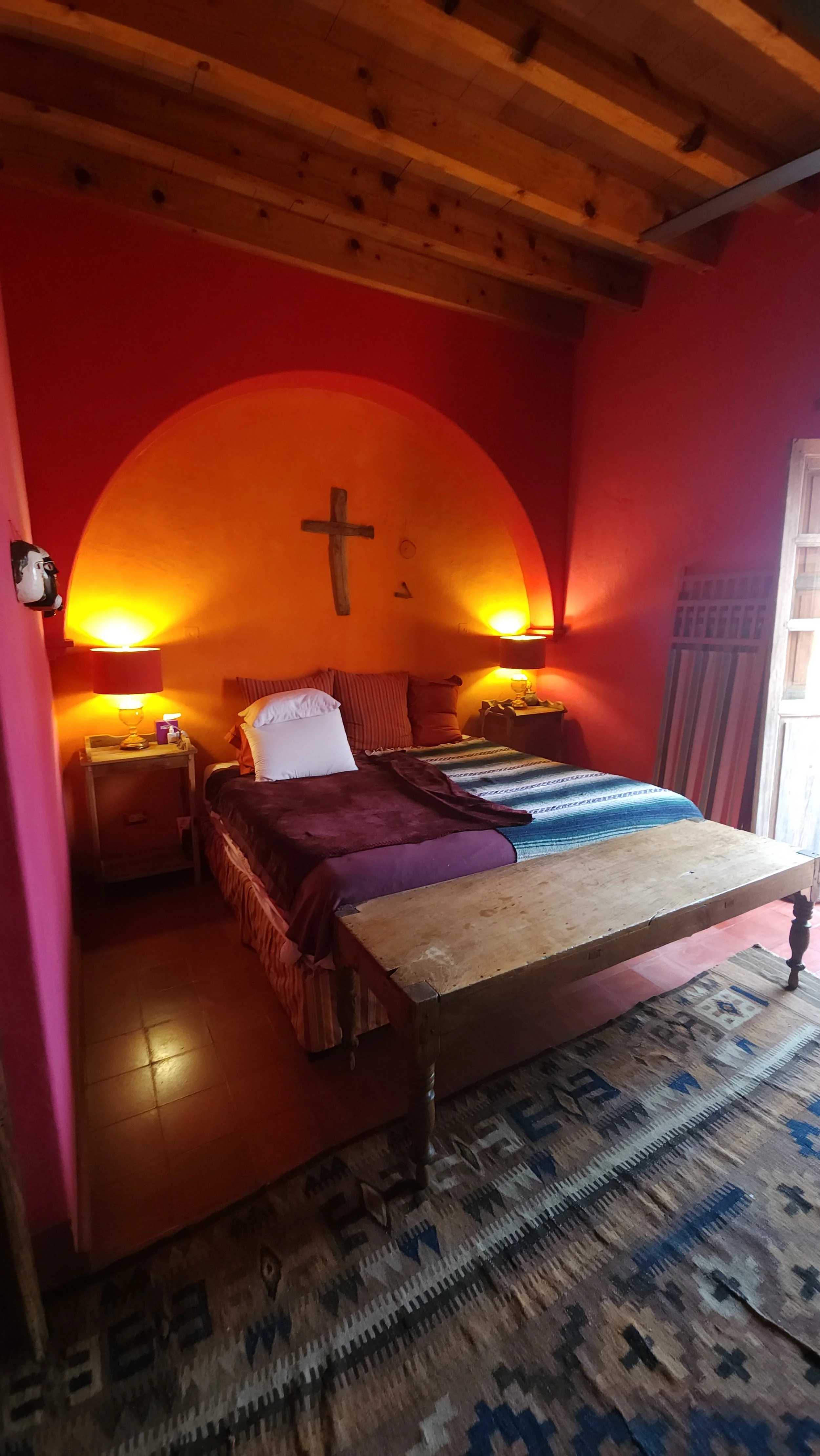 Habitación con paredes color, techo de madera y alfombra con patrón étnico. La cama tiene varias almohadas y edredones, y hay dos mesas de noche con lámparas encendidas. En la pared del fondo hay una cruz de madera y una luz que crea un arco de luz en la pared.