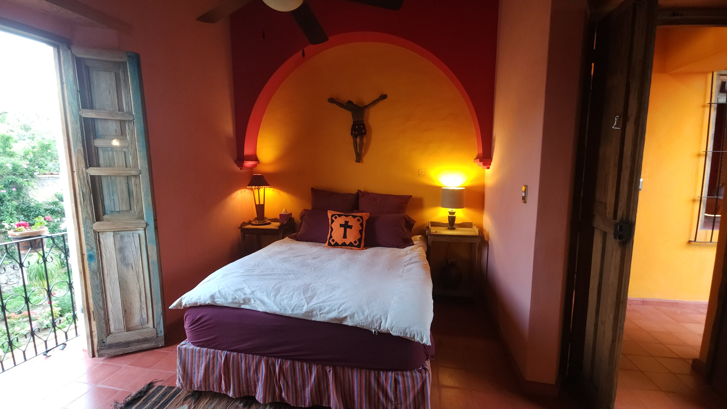 Habitación con cama, almohadas moradas, cortinas, dos lámparas encendidas, crucifijo en la pared y un balcón con vista a plantas y flores.