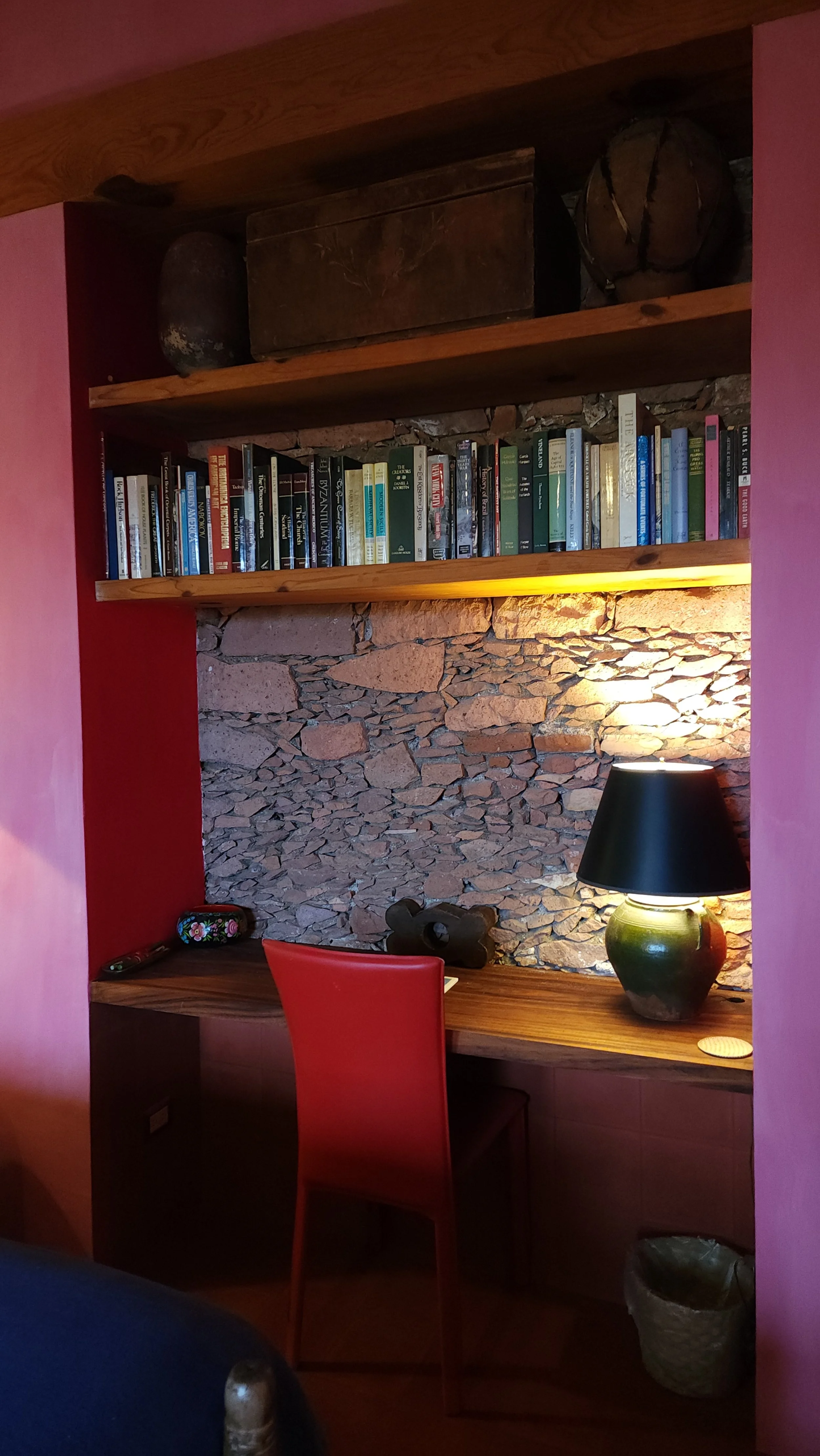 Estudio con paredes de piedra, estantes de madera con libros, escritorio de madera con lámpara y silla roja.