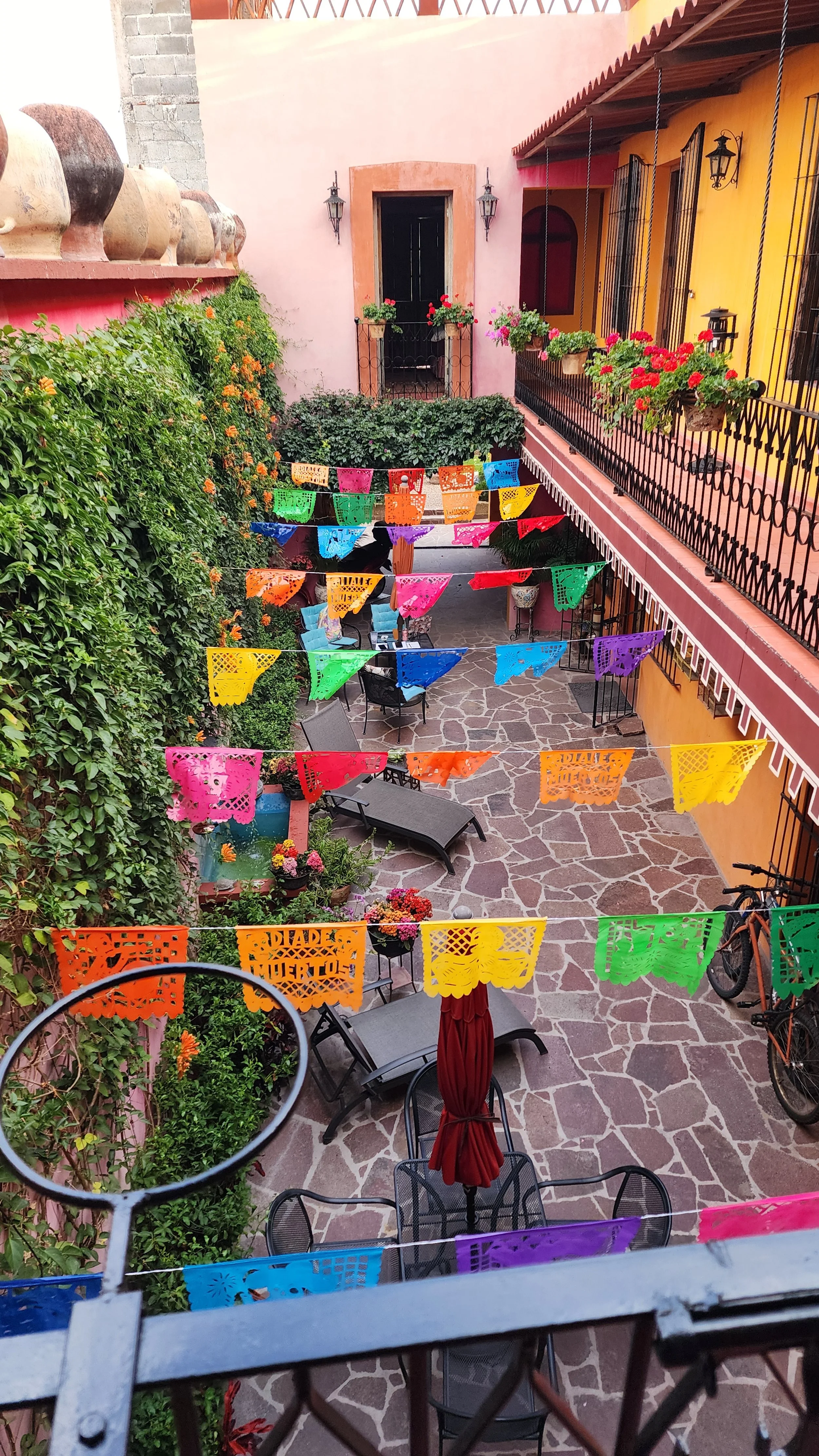 Vista de un patio decorado con banderines de colores colgando en el aire, mesas, sillas, plantas, y paredes pintadas de colores vibrantes.