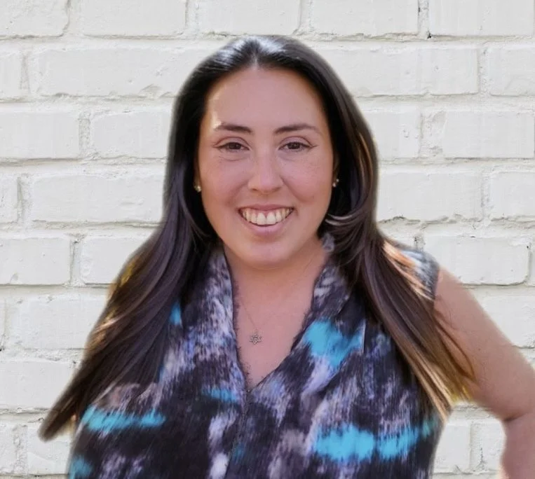 A woman with long dark hair smiling in front of a white brick wall, wearing a sleeveless top with a blue, black, and white abstract pattern and a star necklace.