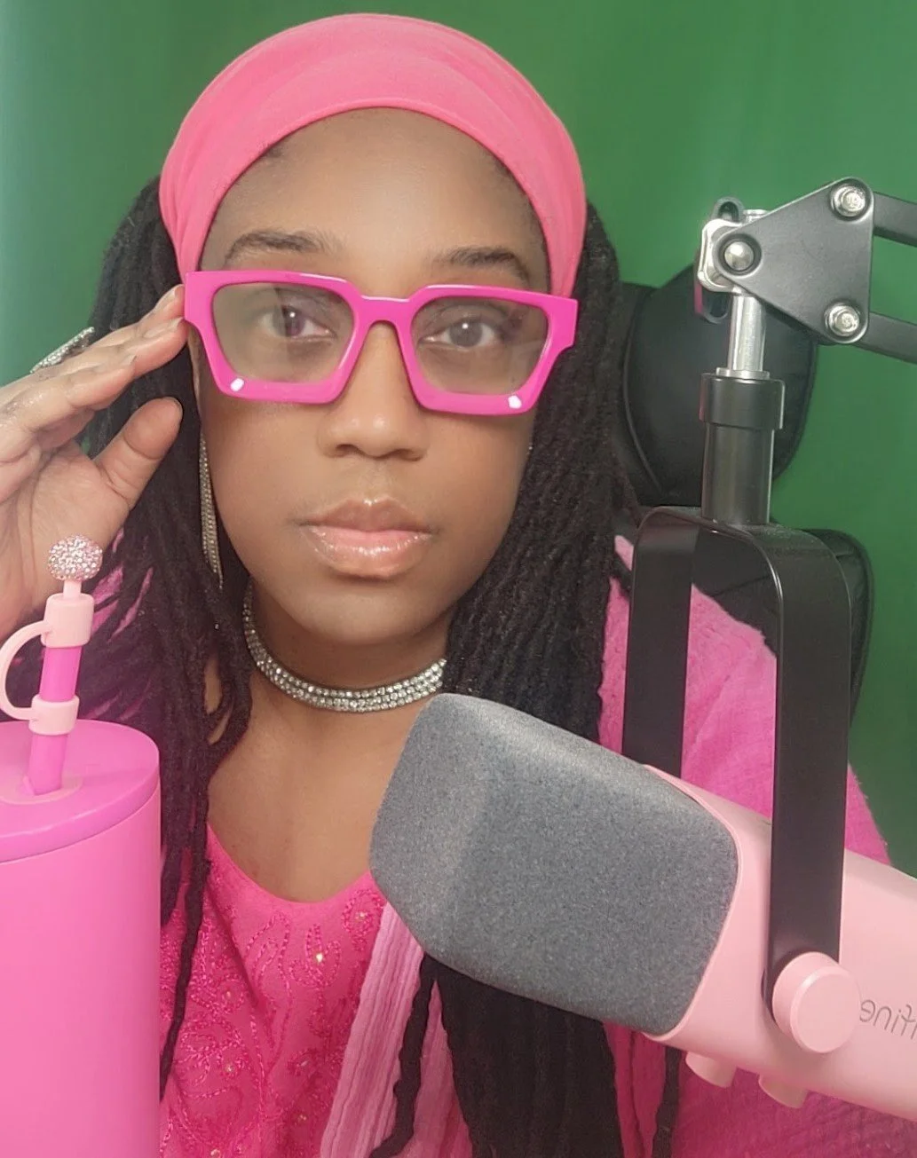 Woman wearing pink glasses, pink headwrap, pink top, and layered necklaces sits in front of a green screen with a microphone and studio equipment.