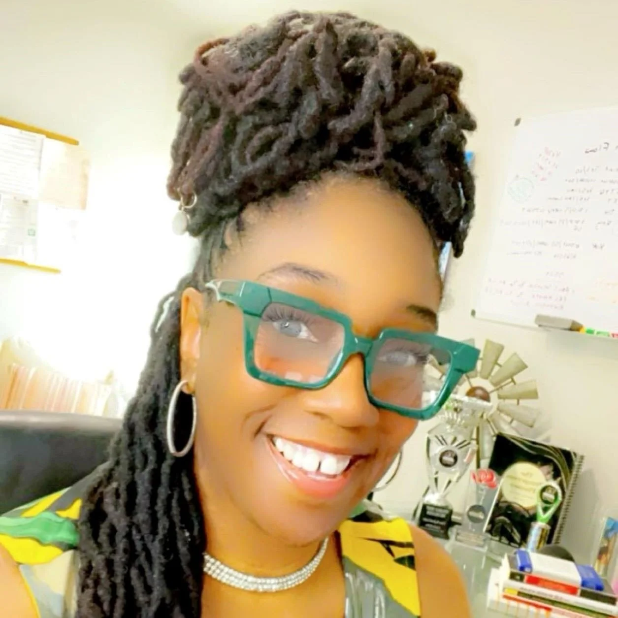 Close-up of smiling woman with styled dreadlocks wearing glasses, hoop earrings, and a yellow top, in an office with whiteboard and trophies in the background.