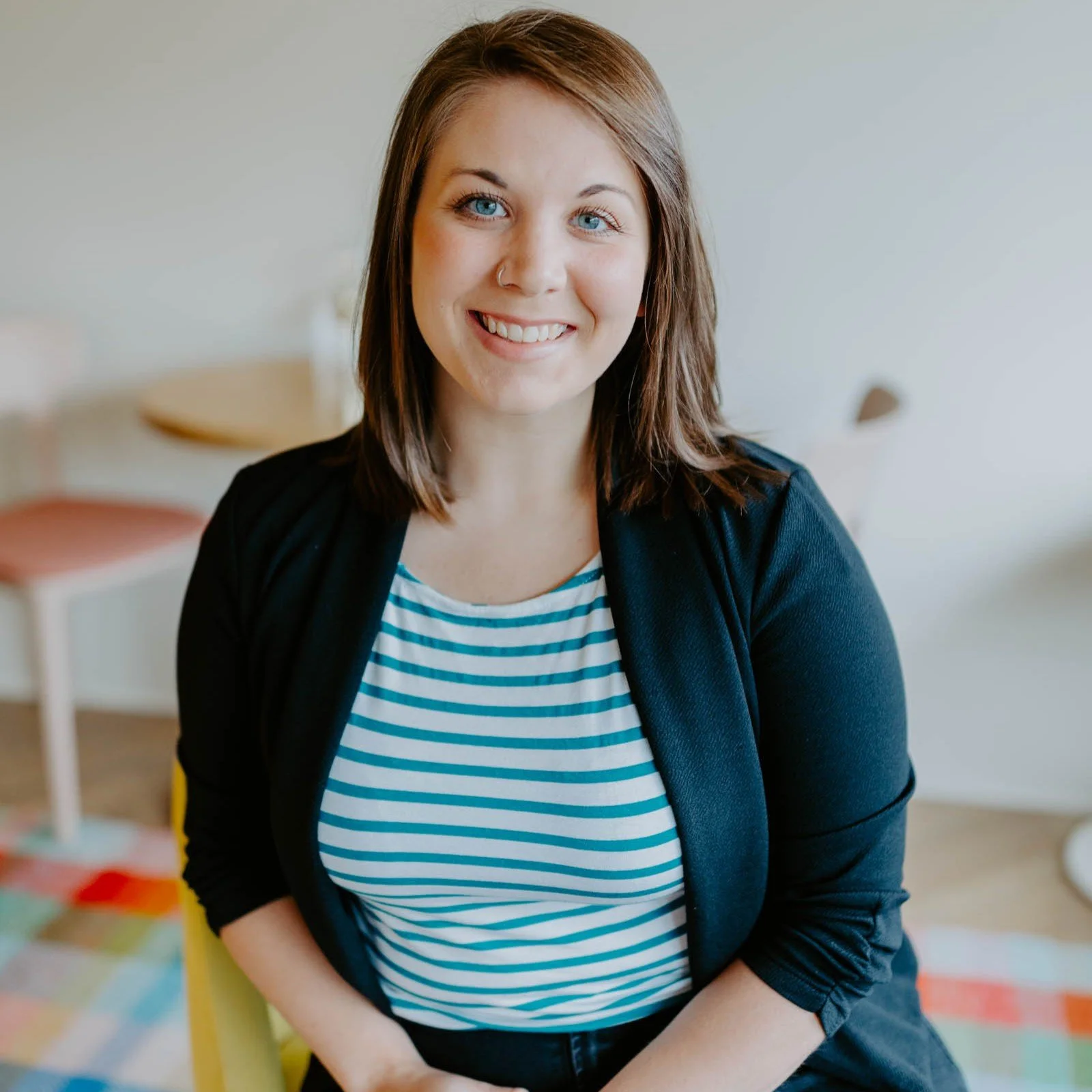 A smiling woman with shoulder-length brown hair, wearing a black blazer over a white and blue striped shirt, sitting in a well-lit room.