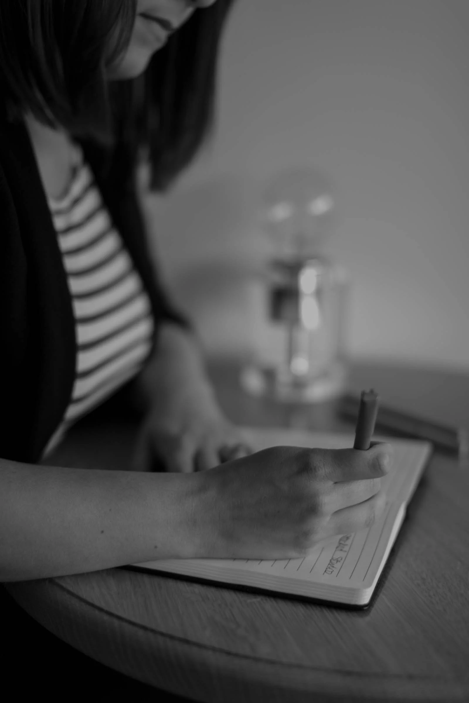 A person writing in a notebook on a wooden table with a lamp in the background.