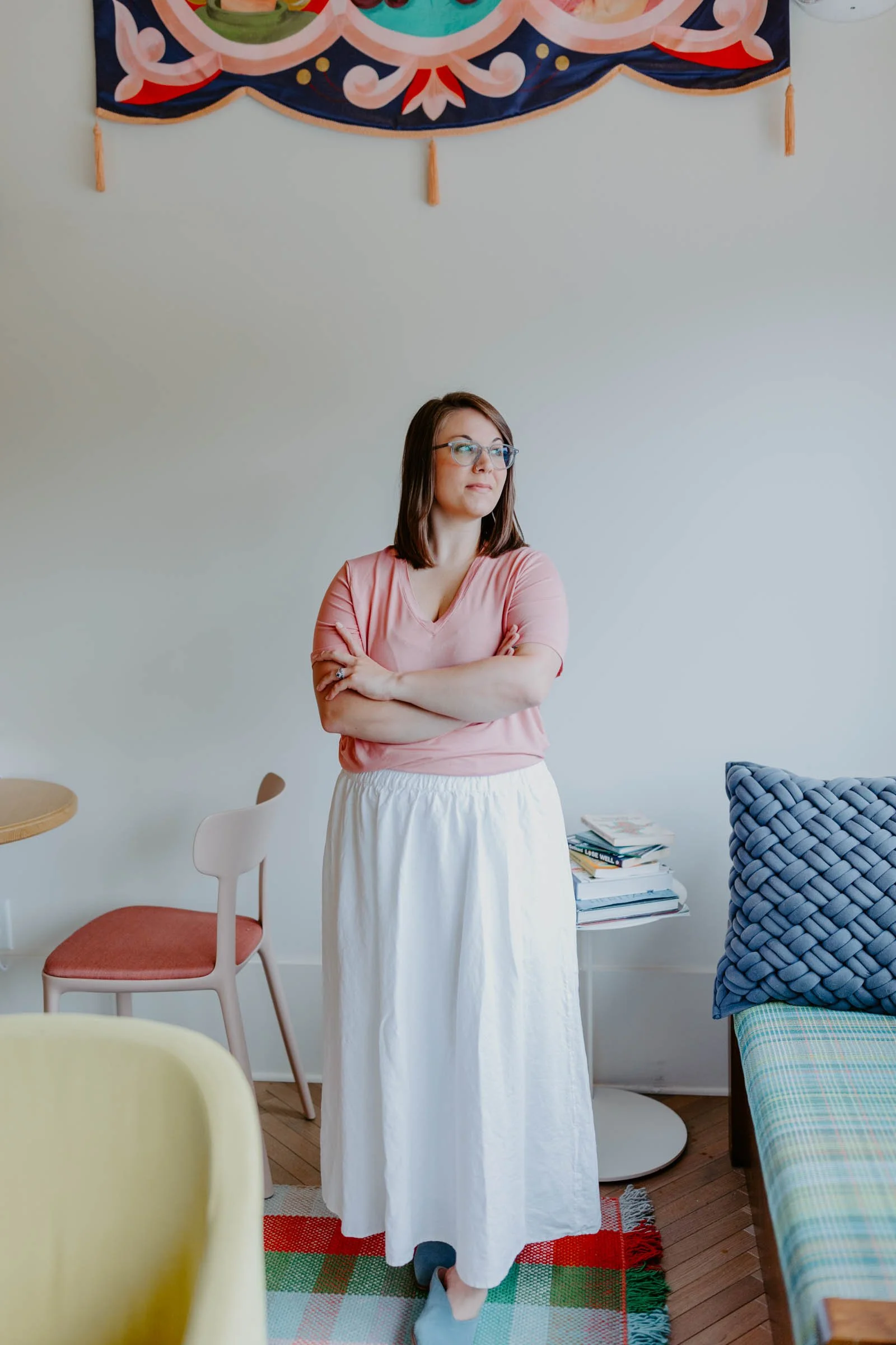 A woman with glasses, wearing a pink top and a white skirt, standing with her arms crossed in a room with a colorful wall hanging and a stack of books on a small table.