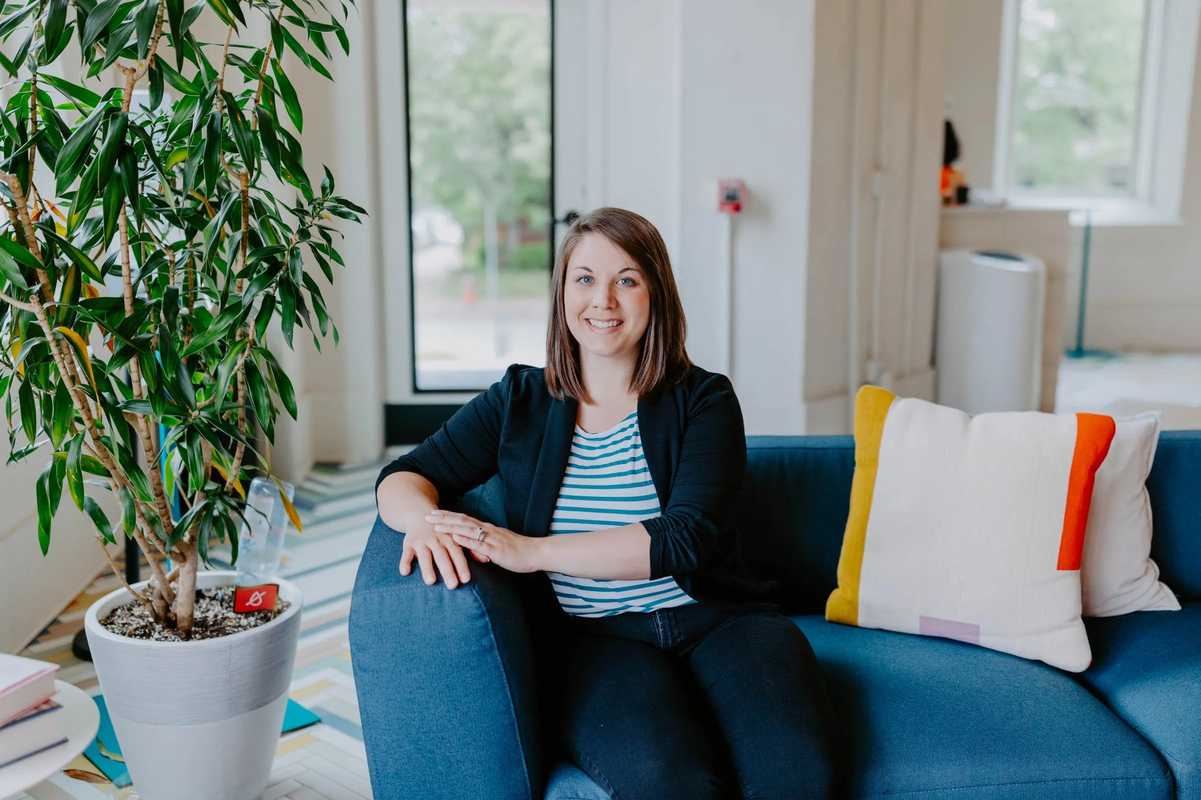 Amanda has shoulder-length brown hair and a black blazer, sitting on a blue couch in a bright room, next to a large potted plant and decorative pillows.