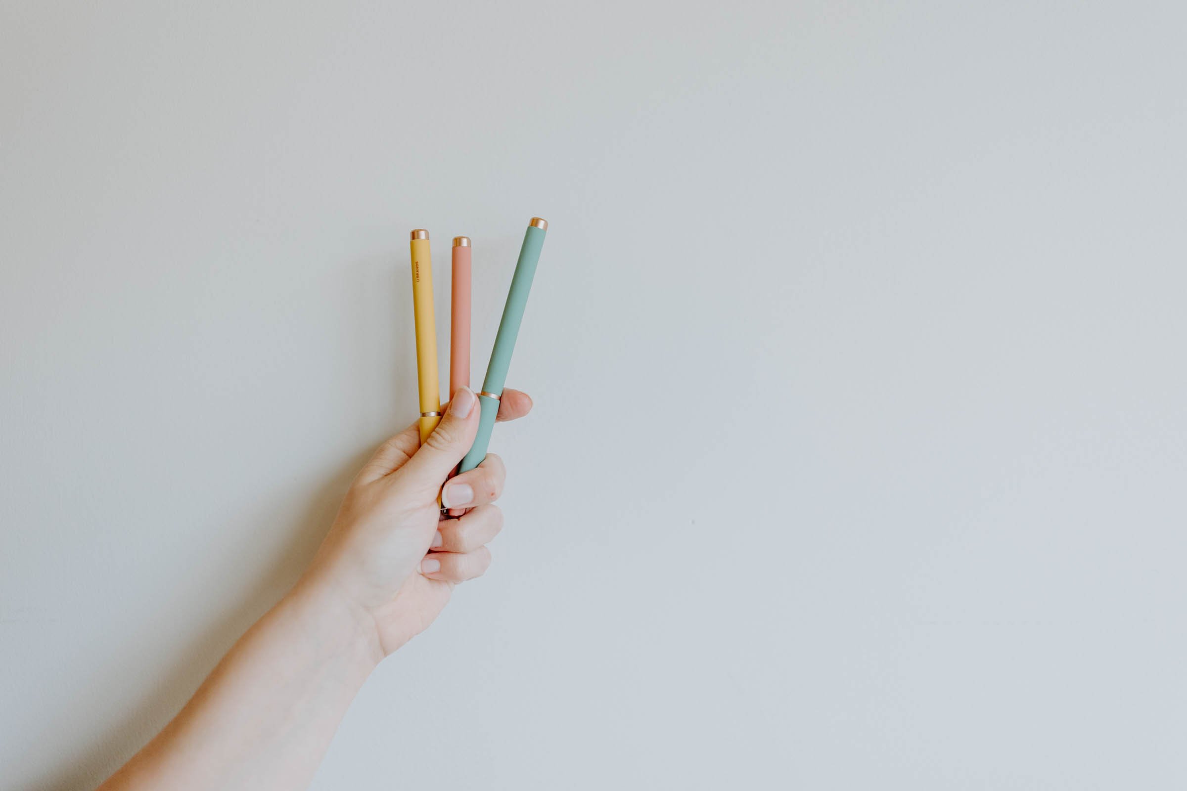 A hand holding three pastel-colored pens against a plain white background.