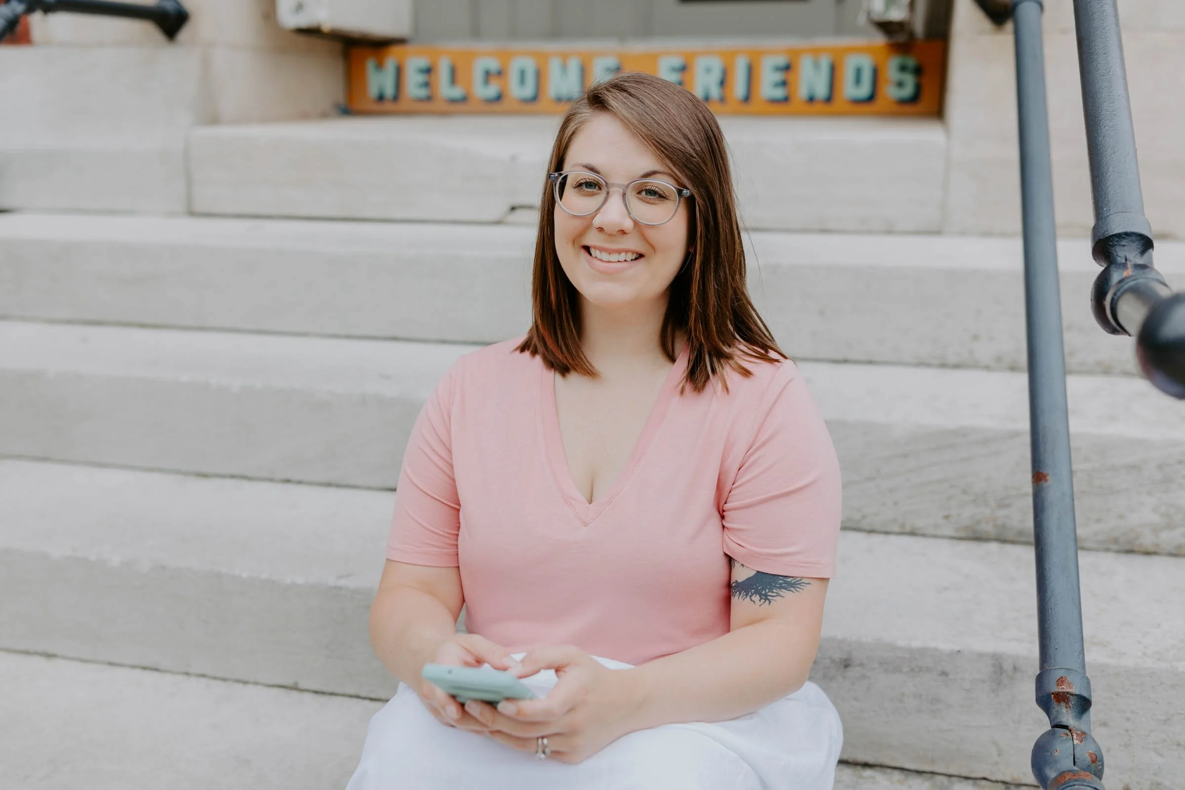A young woman with shoulder-length brown hair, wearing glasses, a pink V-neck T-shirt, and white pants, sitting on outdoor concrete steps, holding a smartphone and smiling. A 'Welcome Friends' sign is visible in the background.