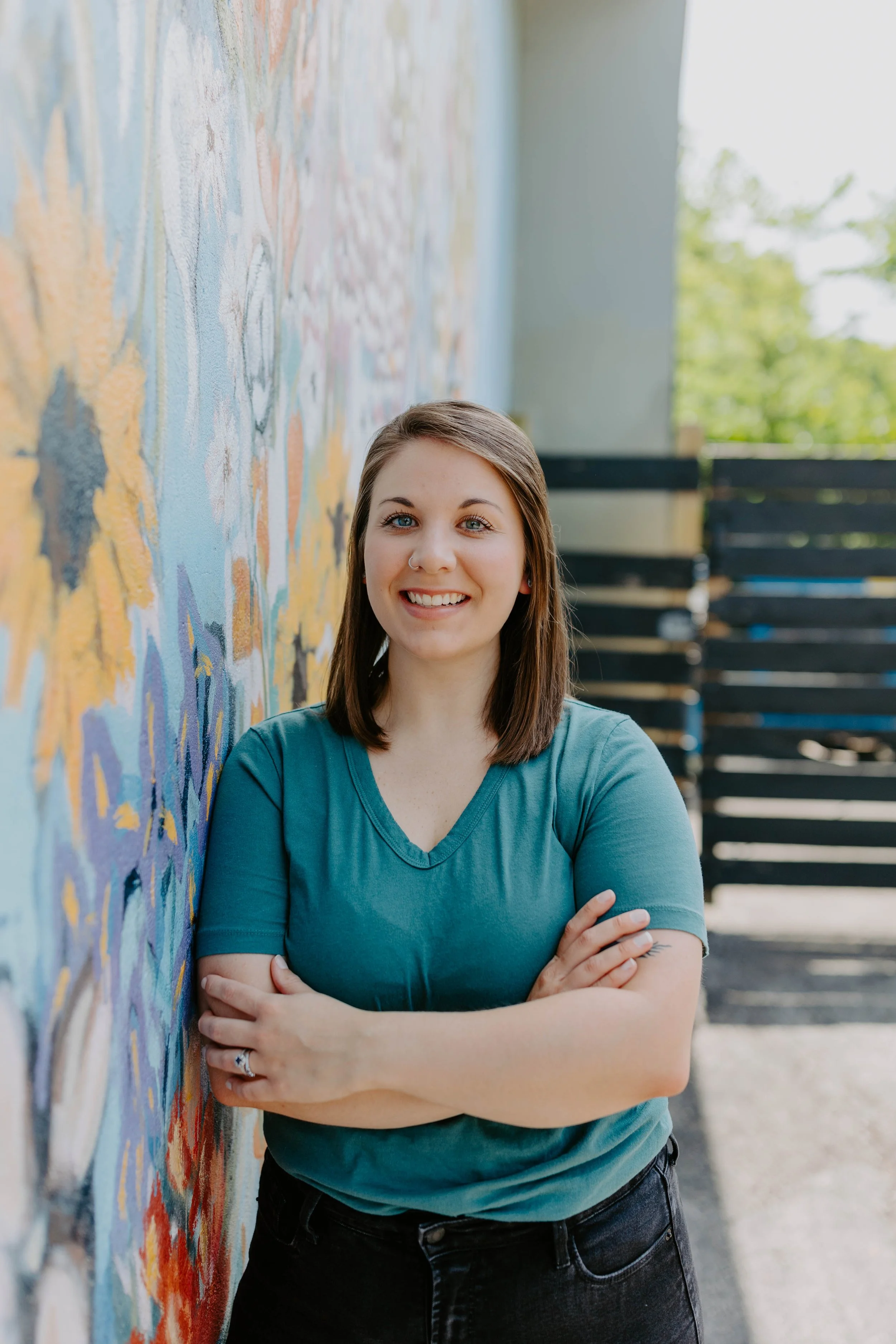 Amanda is a young woman with shoulder-length brown hair, smiling, standing with arms crossed against a colorful mural on a wall outdoors.