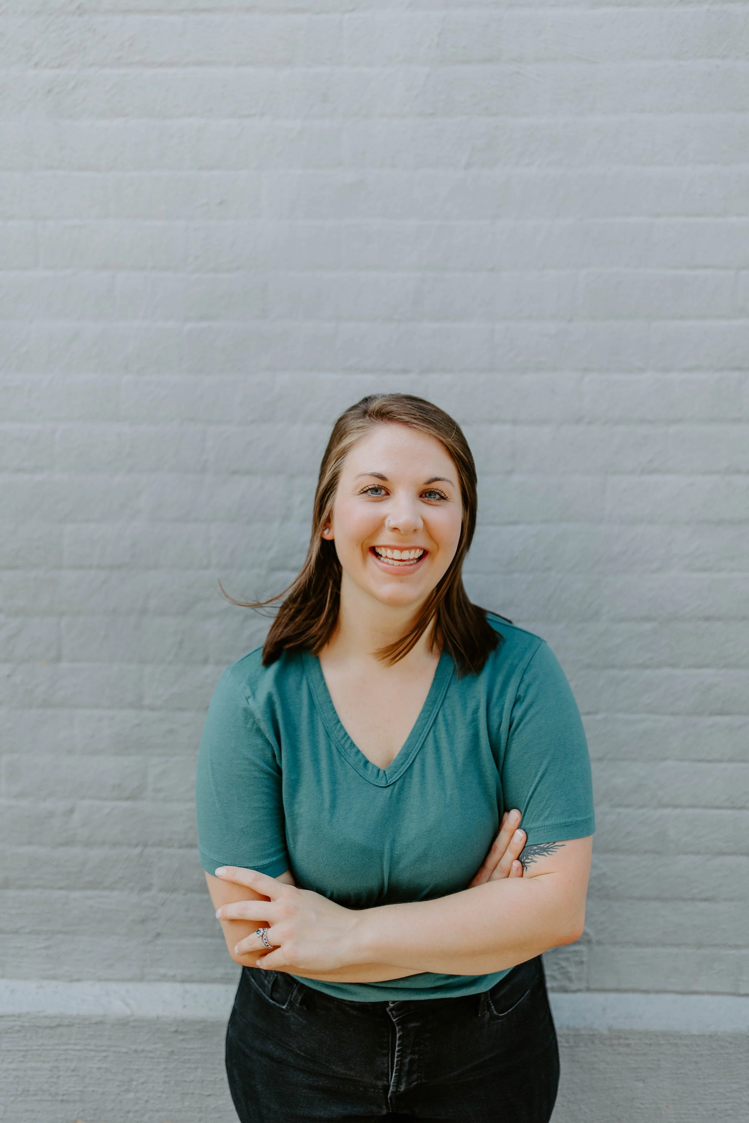 Amanda is a young woman with shoulder-length brown hair and a big smile, wearing a teal t-shirt and black pants, standing with her arms crossed in front of a light gray brick wall.