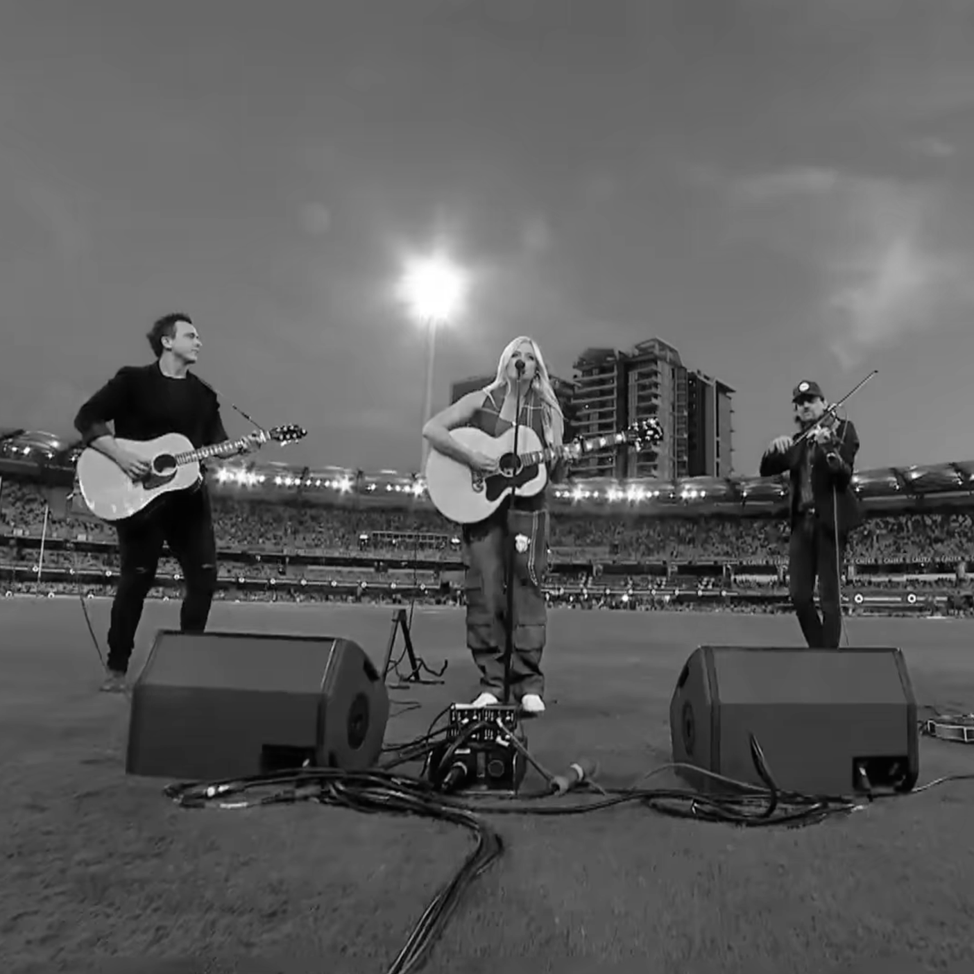 A band performing in a stadium, with three musicians playing guitars and a violin, under an overcast sky with a bright moon or setting sun in the background. Artist include Sara Berki and Luke Moller.