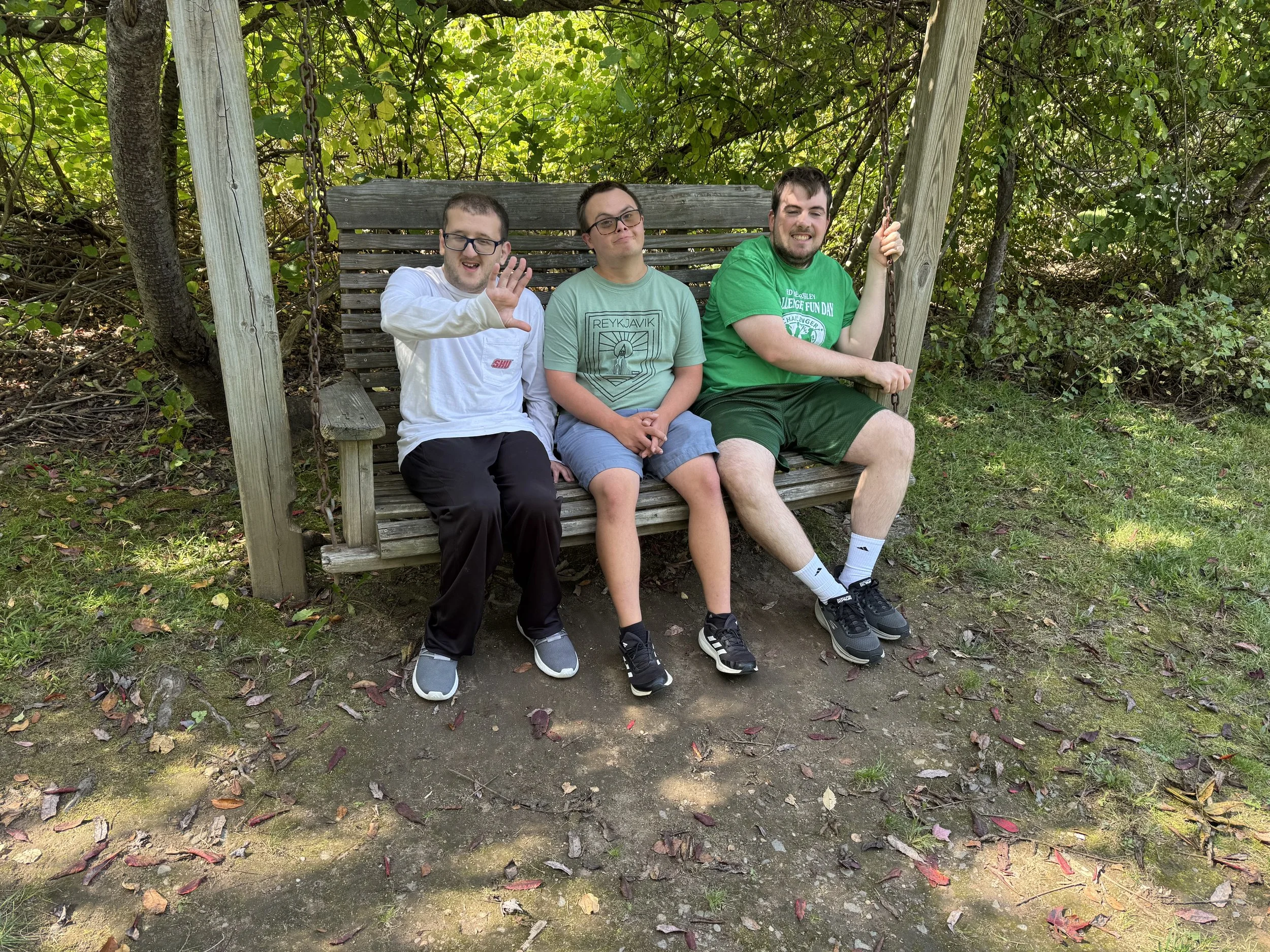 Three young men sitting on a wooden outdoor swing bench surrounded by green foliage. The man on the left is waving and wearing glasses, a white sweatshirt, and black pants. The middle man is wearing a light green T-shirt and shorts, while the man on the right, also wearing glasses, is dressed in a green T-shirt and shorts, holding the swing's pole.