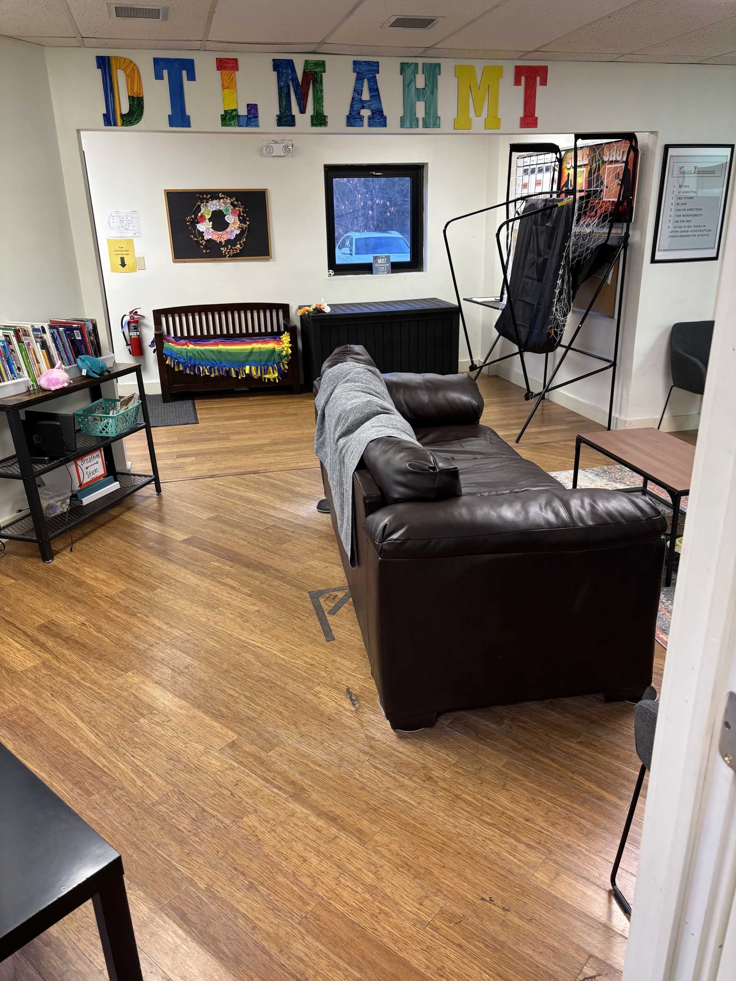 A cozy reading and relaxation room with a brown leather couch, a black metal bookshelf with children's books, a colorful rainbow blanket on a wooden bench, a window with a view of a parking lot.