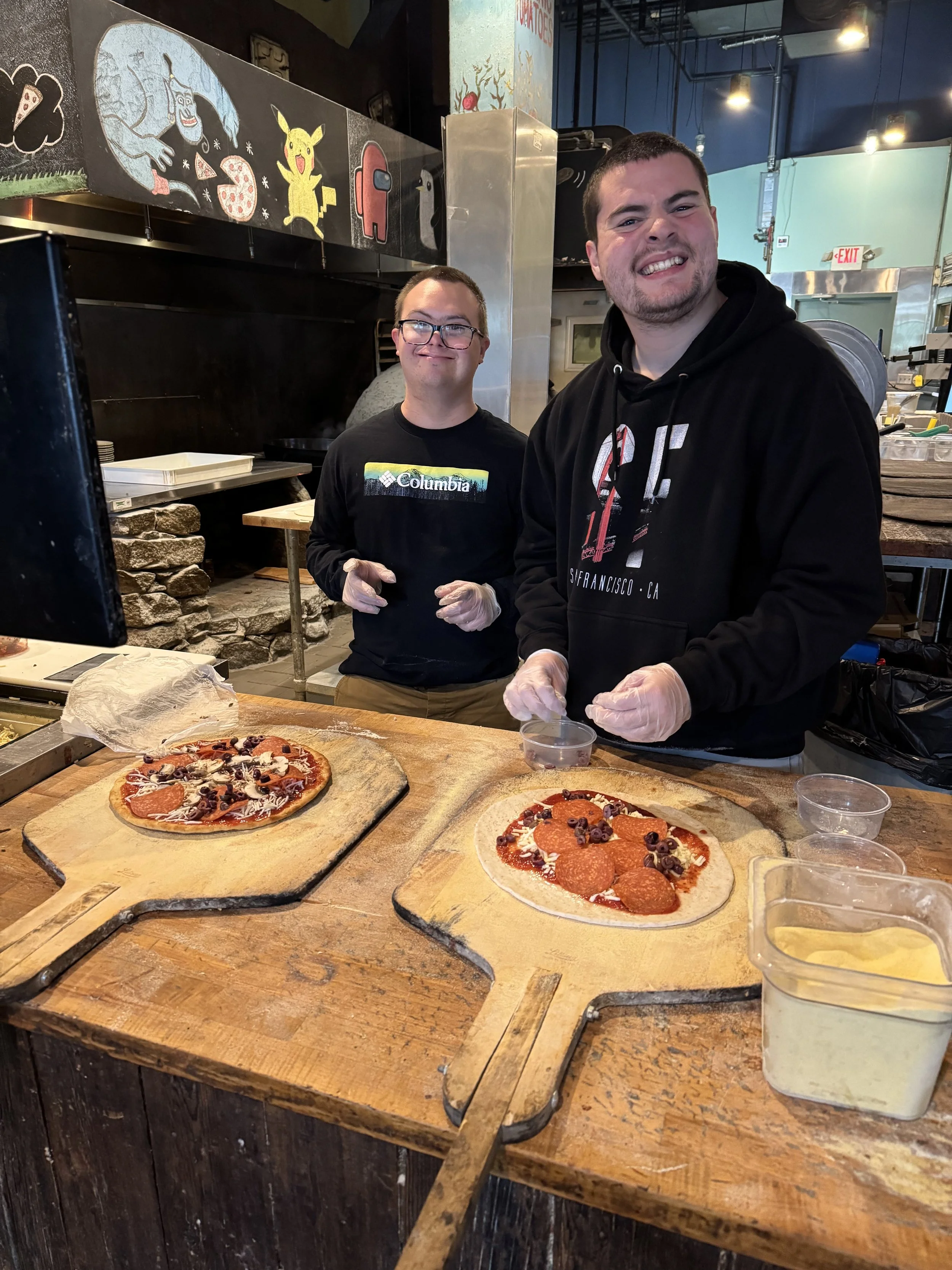 Two men preparing pizzas at a restaurant counter; one is smiling at the camera and the other is standing behind him with a neutral expression. The pizzas are topped with pepperoni, black olives, and shredded cheese.