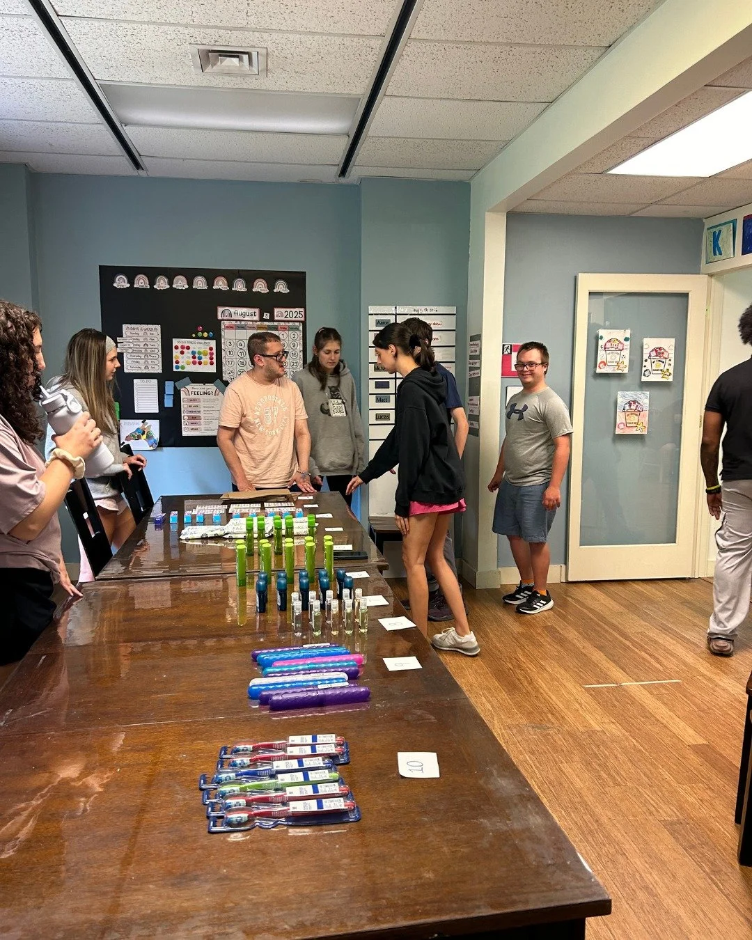 Individuals and staff in a classroom around a table displaying colorful science or art projects, with posters and charts on the wall.