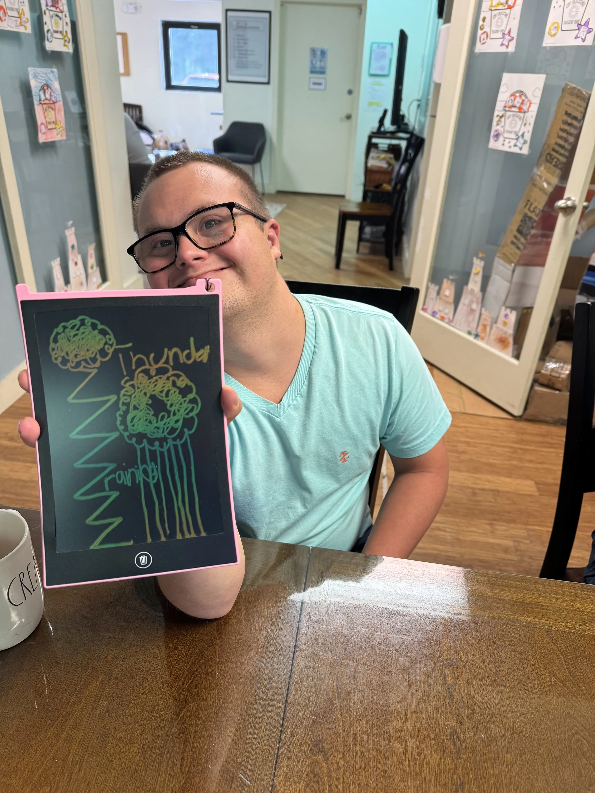Young man with glasses holding a chalkboard sign with colorful scribbles and writing, sitting at a wooden table in a room with decorated walls.