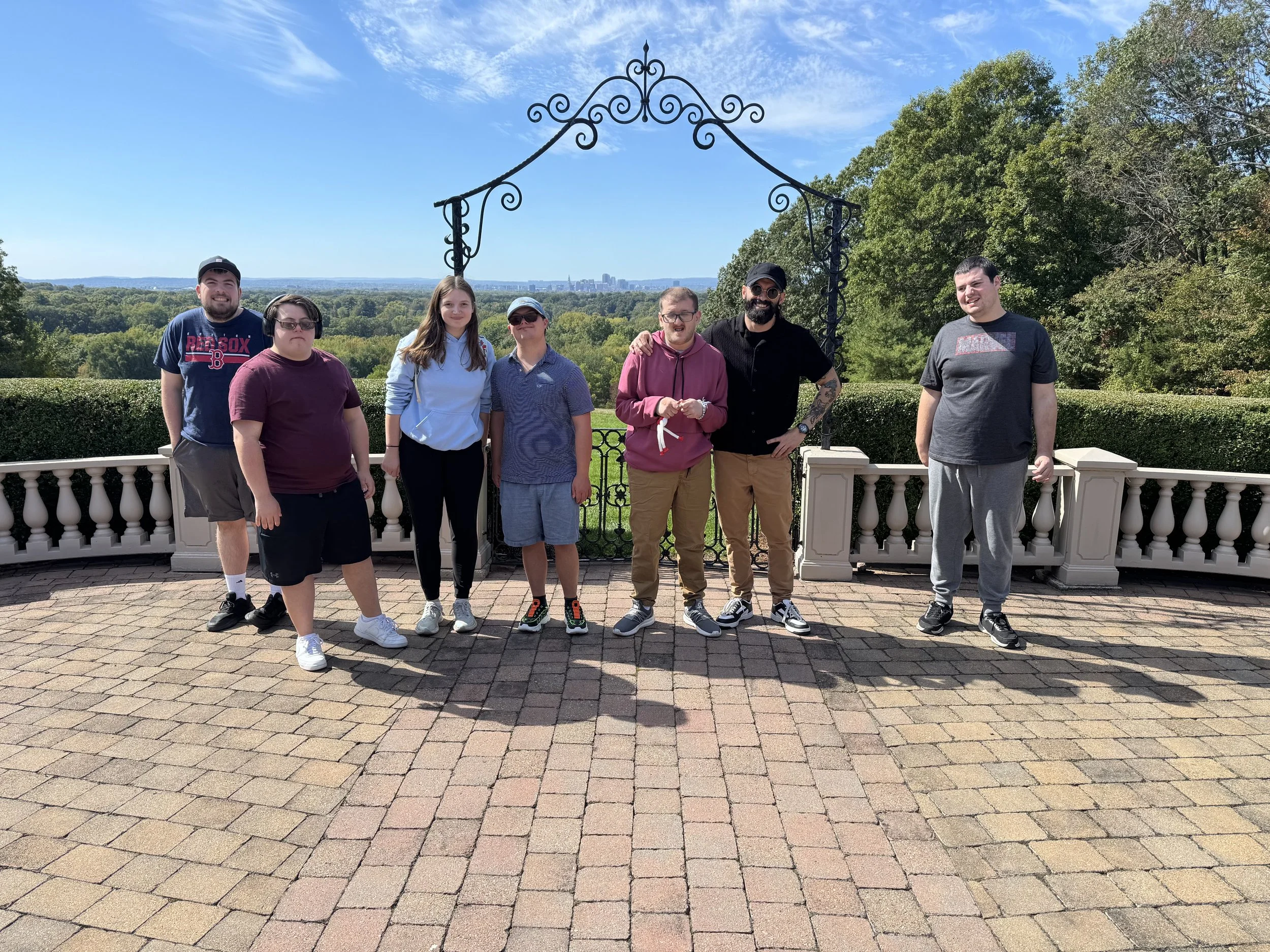 Group of seven people standing outdoors on a brick patio under a decorative metal arch, with a scenic view of trees and distant city skyline in the background on a sunny day.