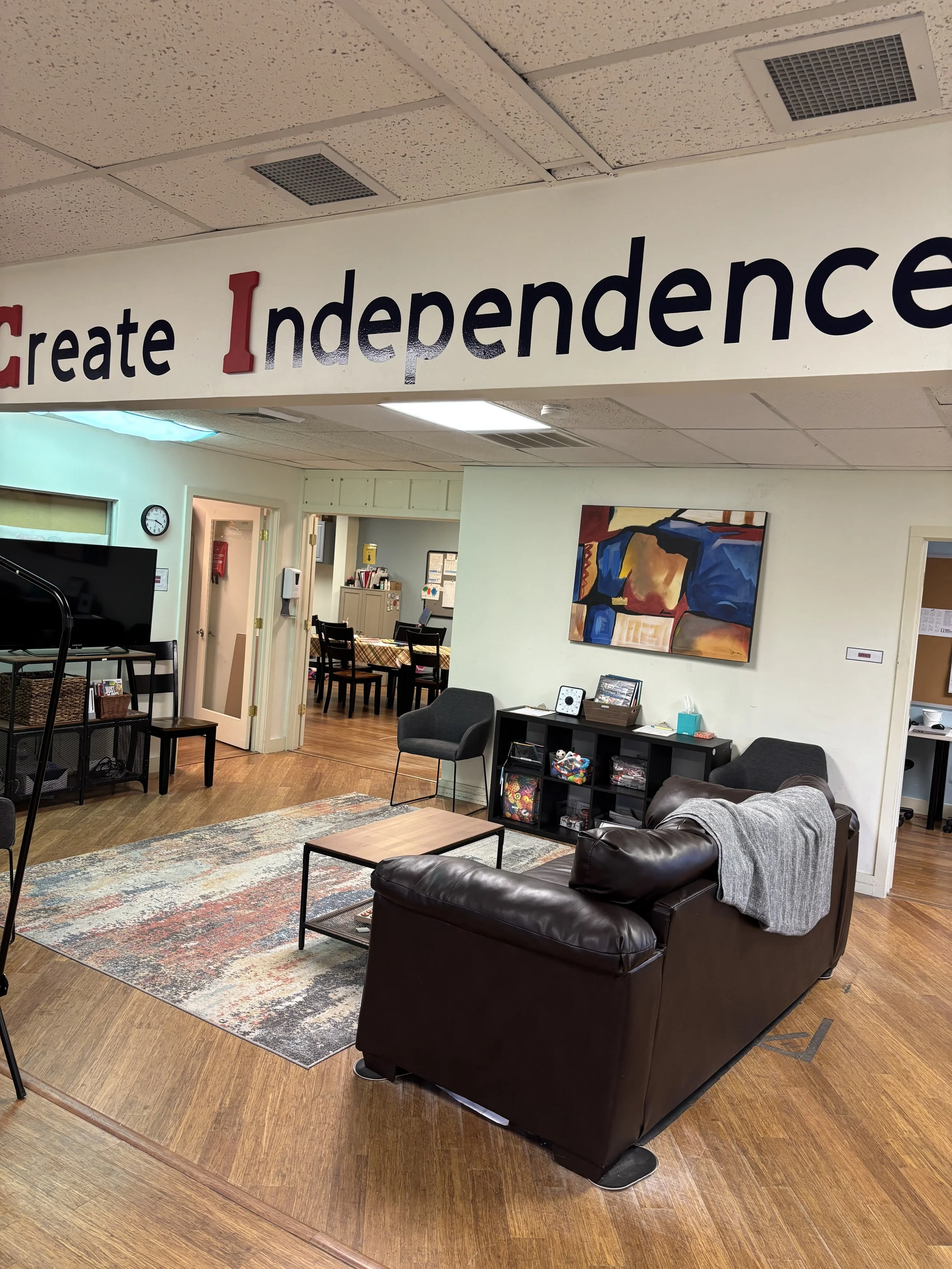 A cozy living room with a leather sofa, two black chairs, a wooden coffee table, a colorful abstract painting on the wall, and a black storage unit filled with books and decorative items. The space has wooden flooring and a patterned area rug. Part o