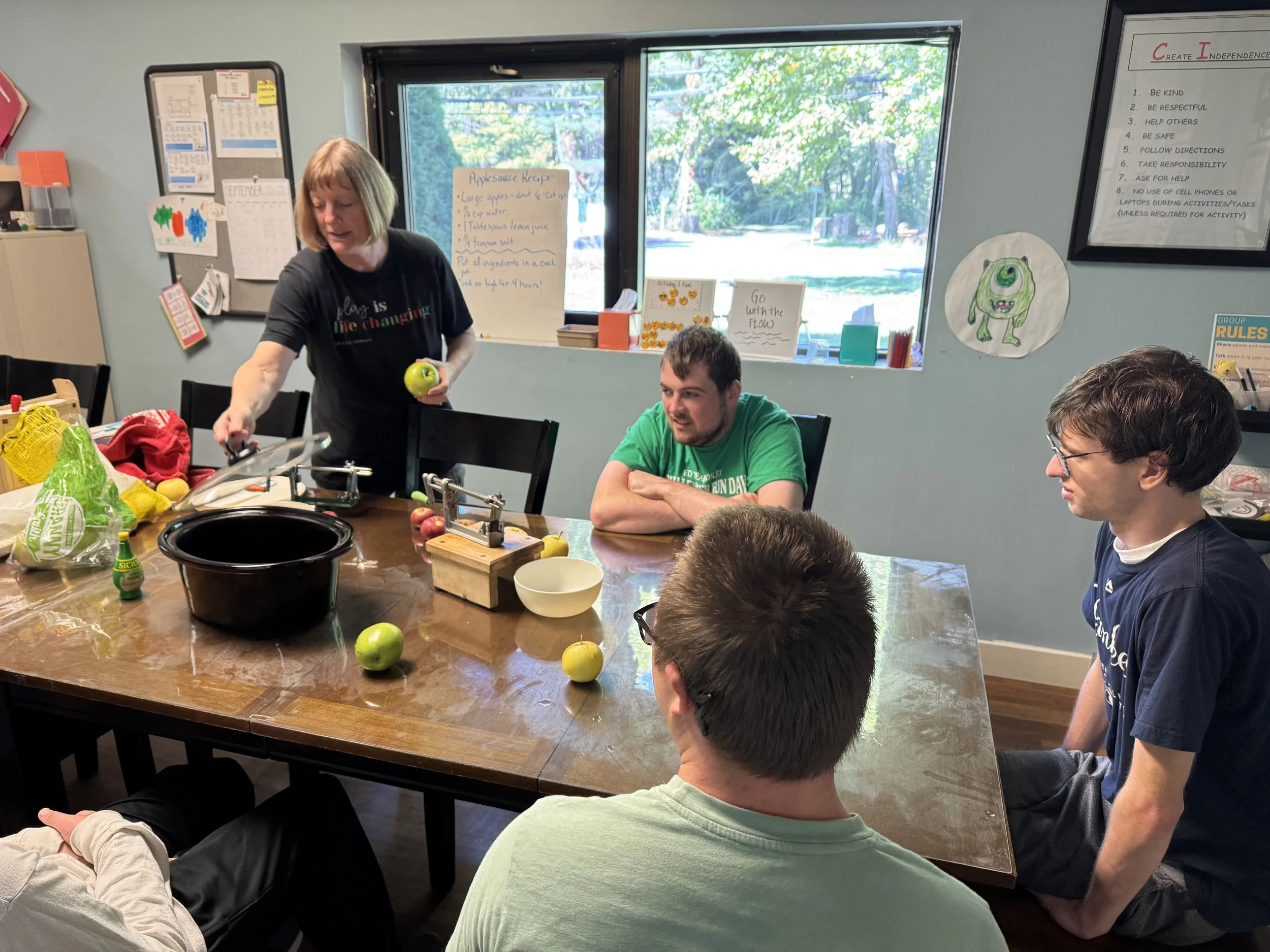 A woman demonstrates apple peeling for a group of four men seated at a wooden table in a room with light blue walls. The table has apples, a black bowl, and some kitchen tools on it. There are windows showing green trees outside, and various posters and drawings are on the walls.