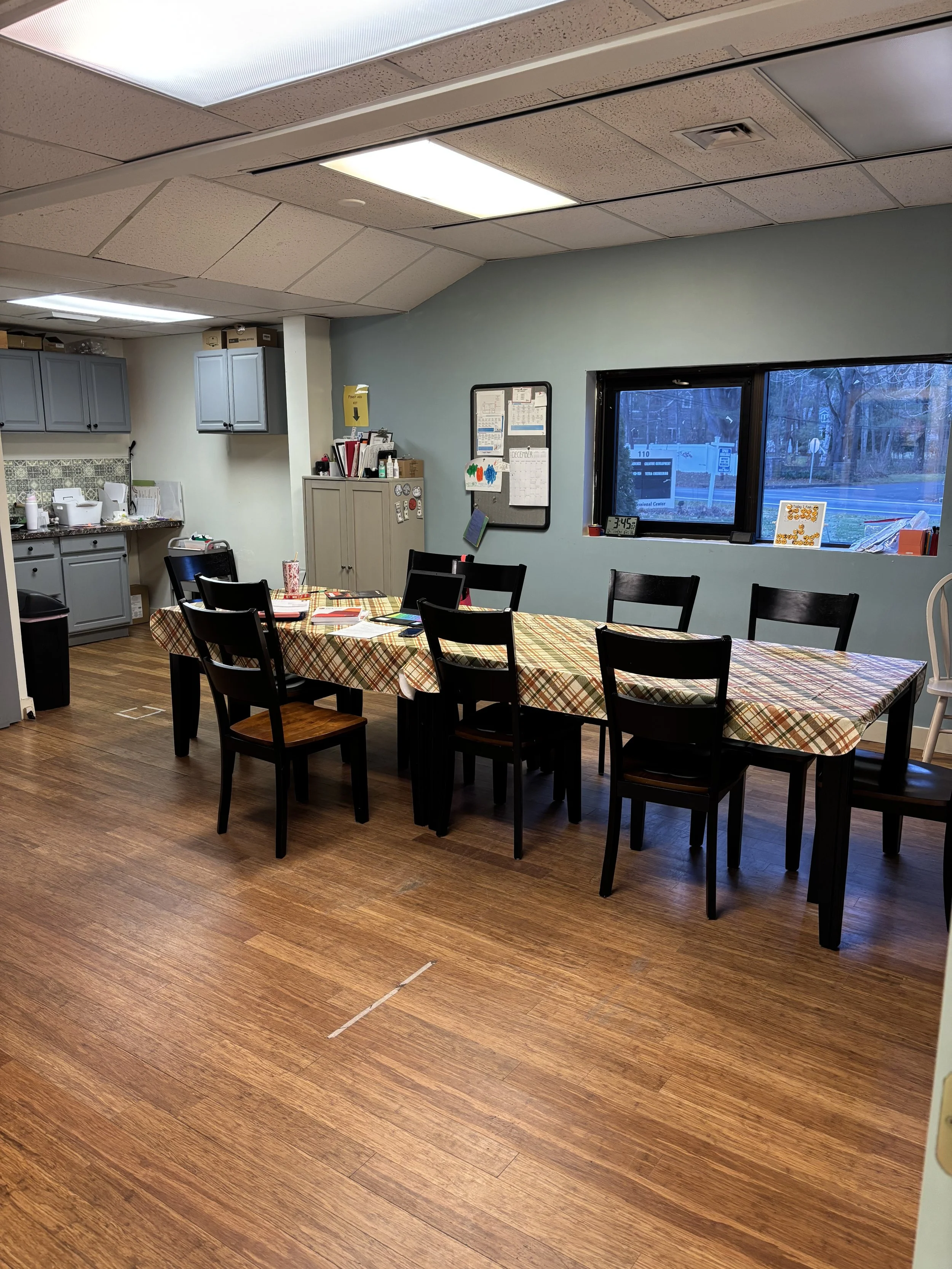 A dining room with wooden flooring and a long table covered with a plaid tablecloth, surrounded by six black chairs. There is a window on the right side showing a street view. In the background, there are kitchen cabinets, a countertop, and storage c