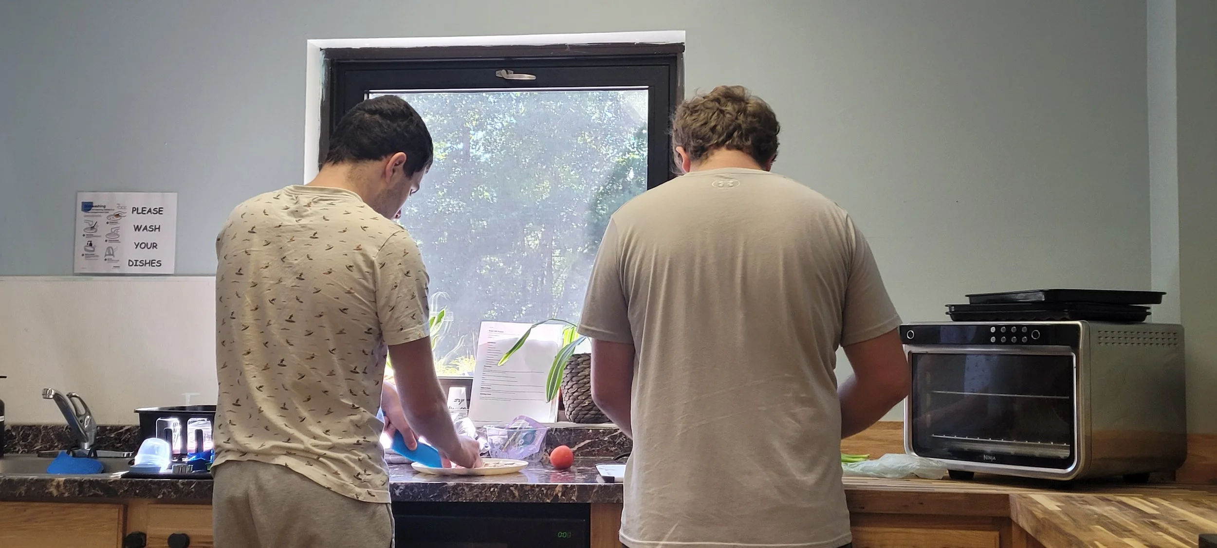 Two young men are washing dishes at a kitchen sink near a window, with various kitchen items on the counter and a sign on the wall that says 'Please wash your dishes'.