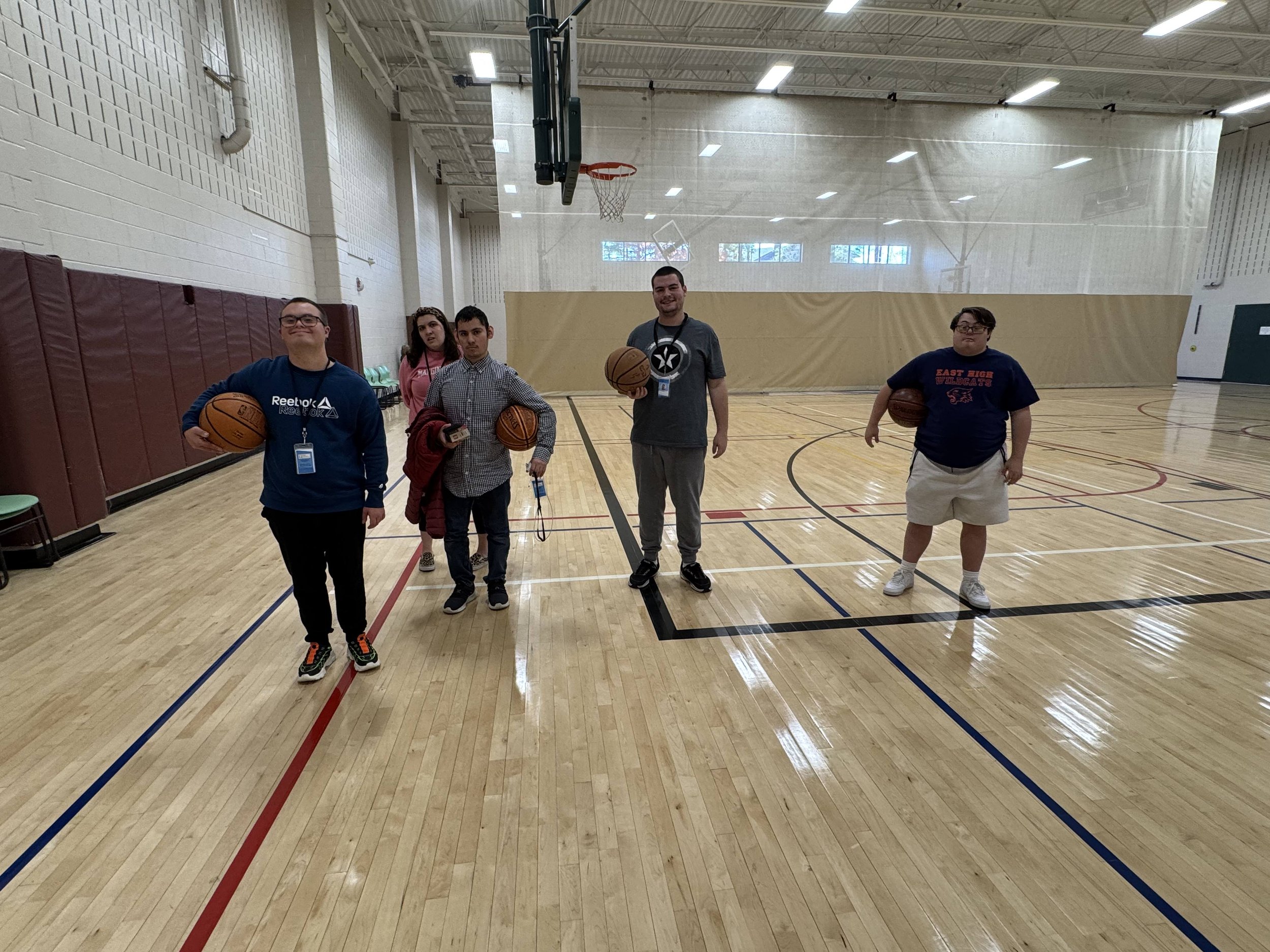 Five teenagers in a gymnasium holding basketballs, standing in a line. The gym has a hardwood floor, basketball hoop, and high ceiling with lights and windows.