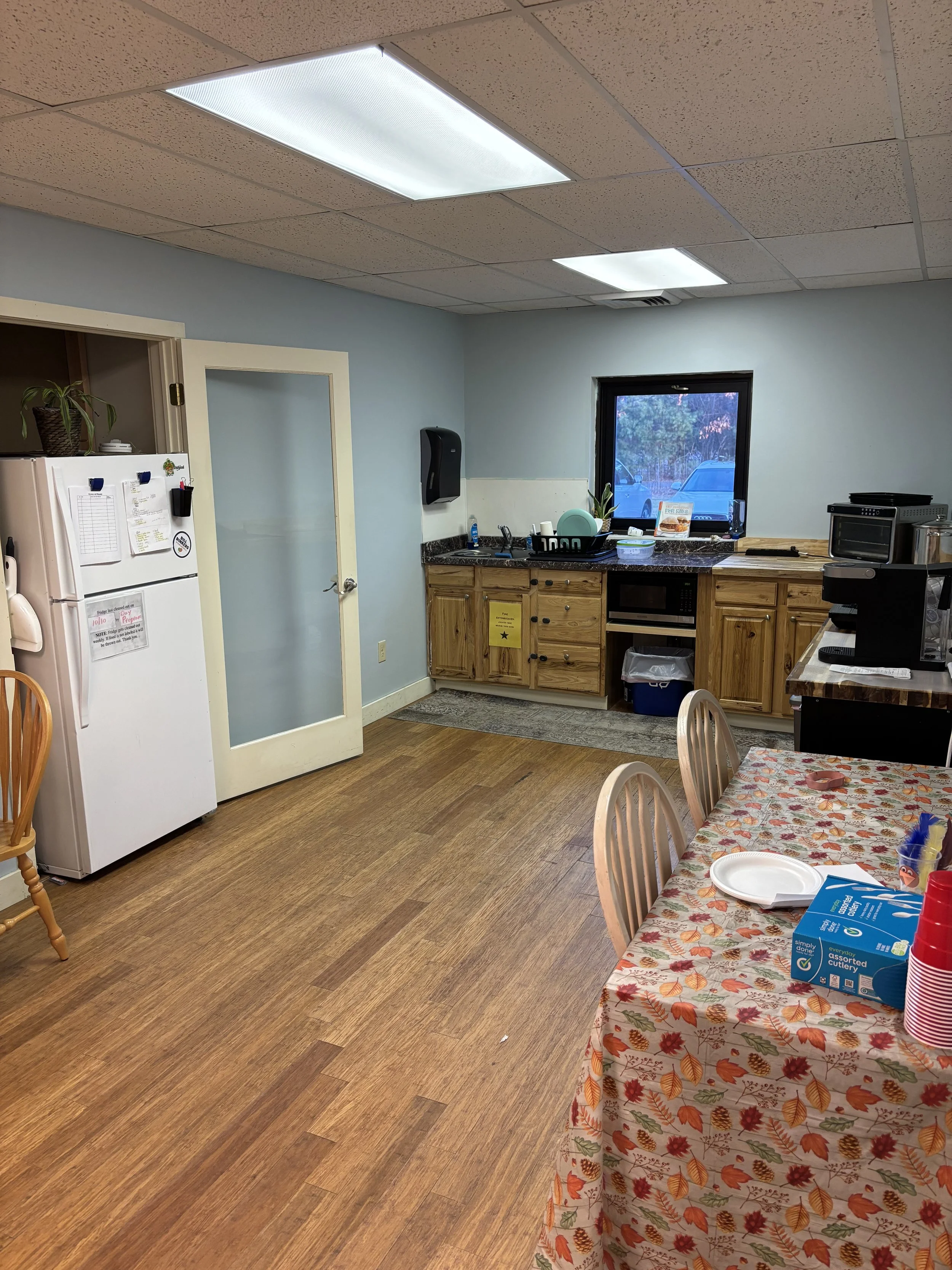 A kitchen with a table covered in a fall-themed tablecloth, a white refrigerator, wooden cabinets, a small window, and various kitchen appliances.