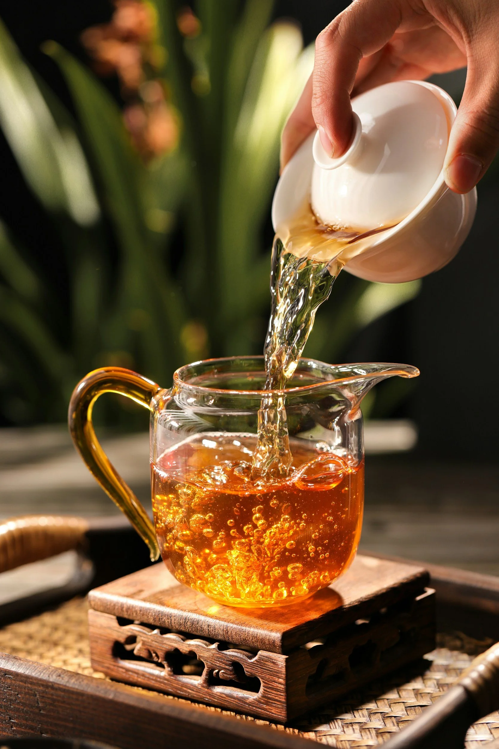 A hand pouring hot tea from a white teapot into a glass cup on a wooden coaster.