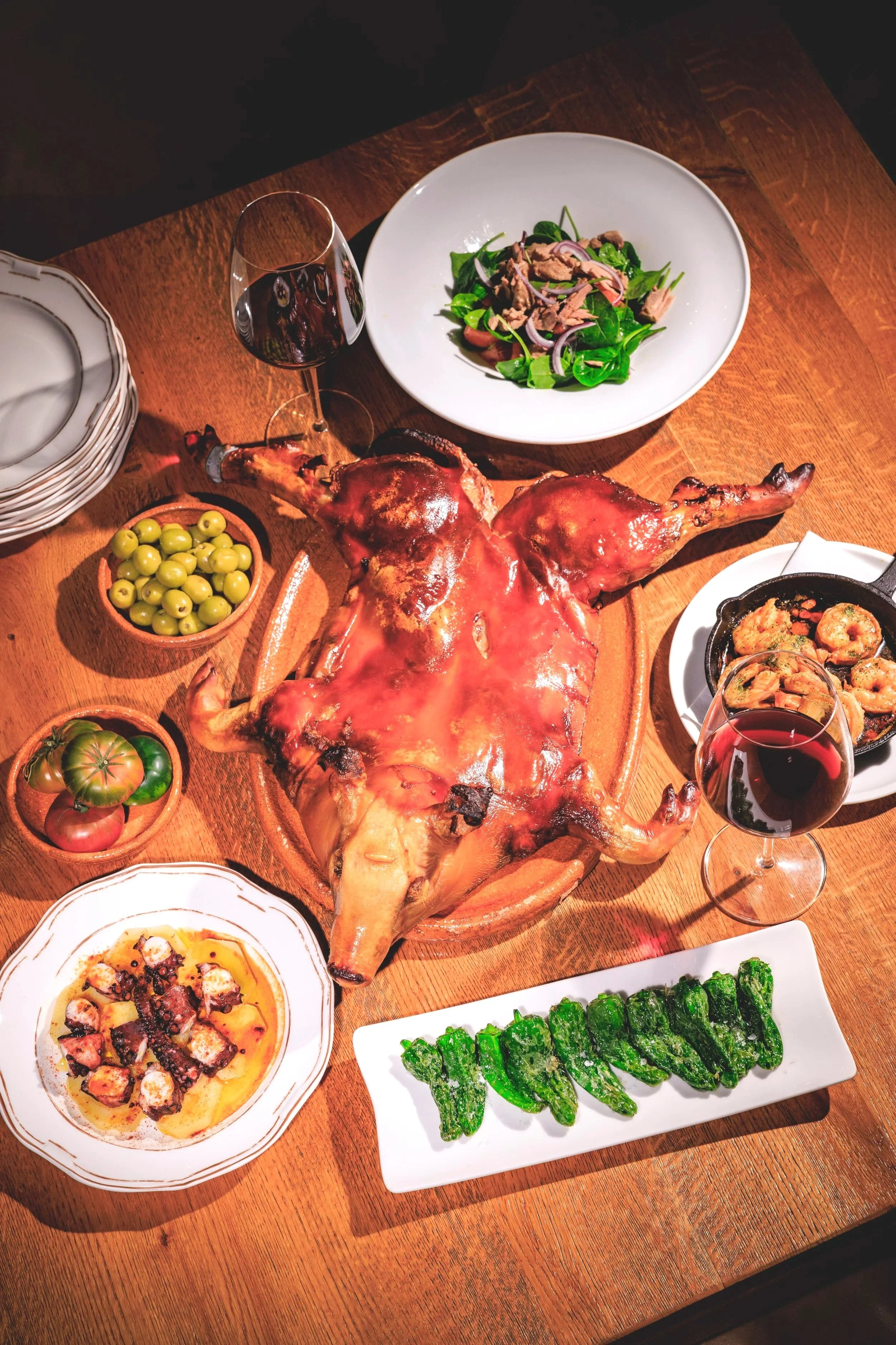 A dinner table with a roasted pig as the centerpiece, surrounded by various dishes including green vegetables, a salad, shrimp, octopus, and drinks like red wine.