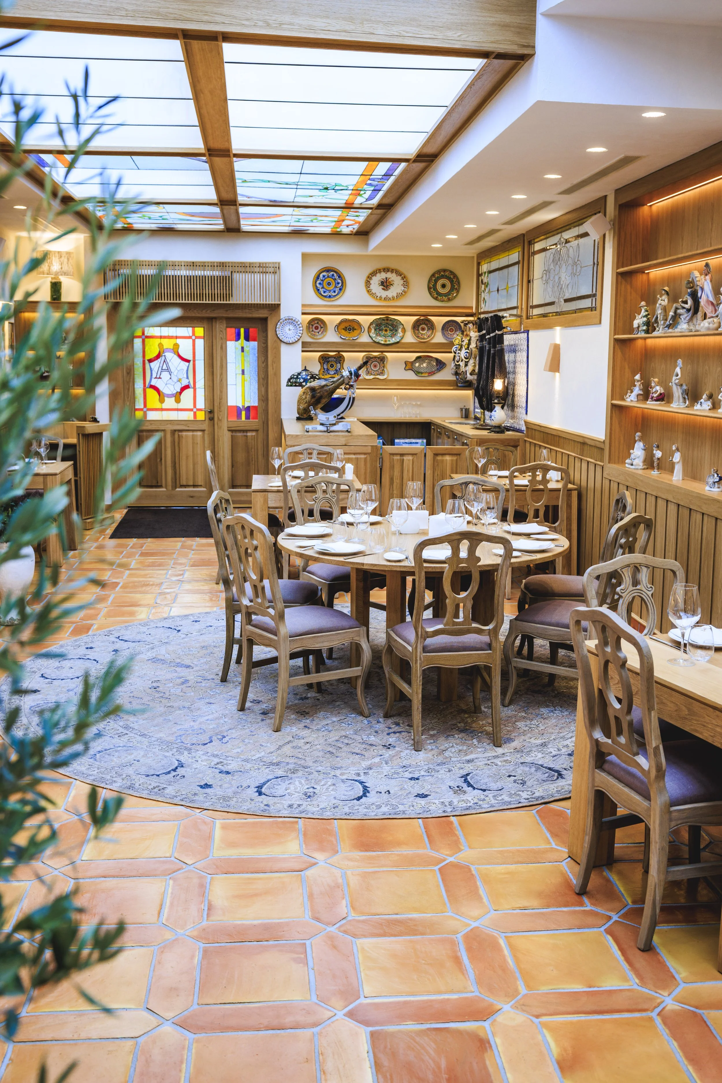 A cozy Italian restaurant with a round dining table set with wine glasses, plates, and napkins, surrounded by wooden chairs with purple cushions. The room features terracotta tile floors, a blue patterned rug, decorative plates on the wall, a stained glass door with a shield emblem, and a wooden display shelf with figurines. A large skylight provides natural light.
