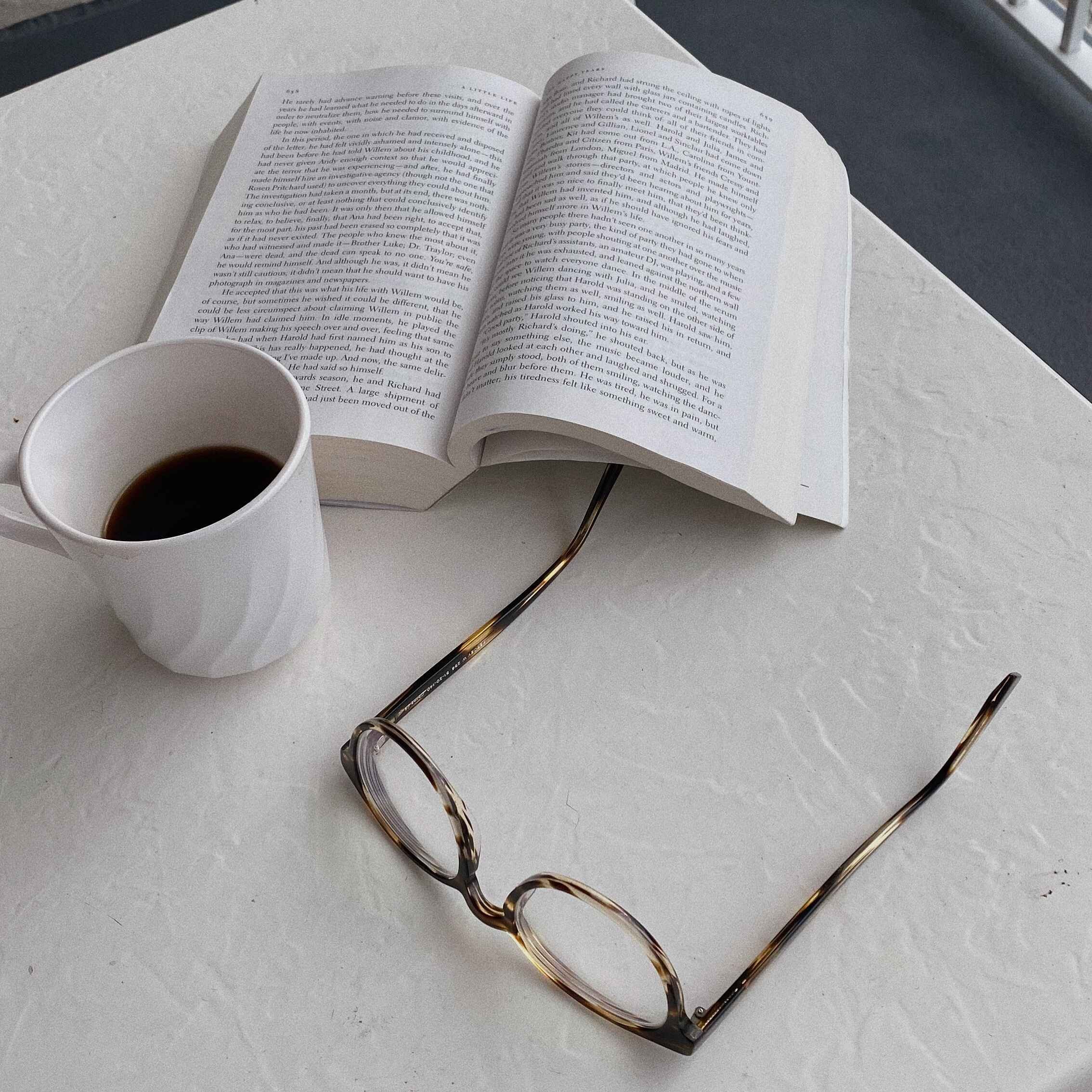 An open book, a white mug filled with black coffee, and a pair of glasses on a white textured table.