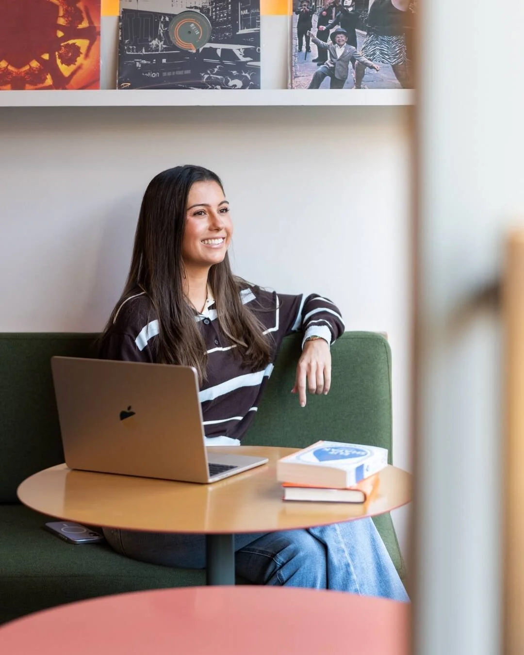 A young woman with long dark hair smiling while sitting on a green couch in a cafe, with a silver MacBook and books on a round wooden table.