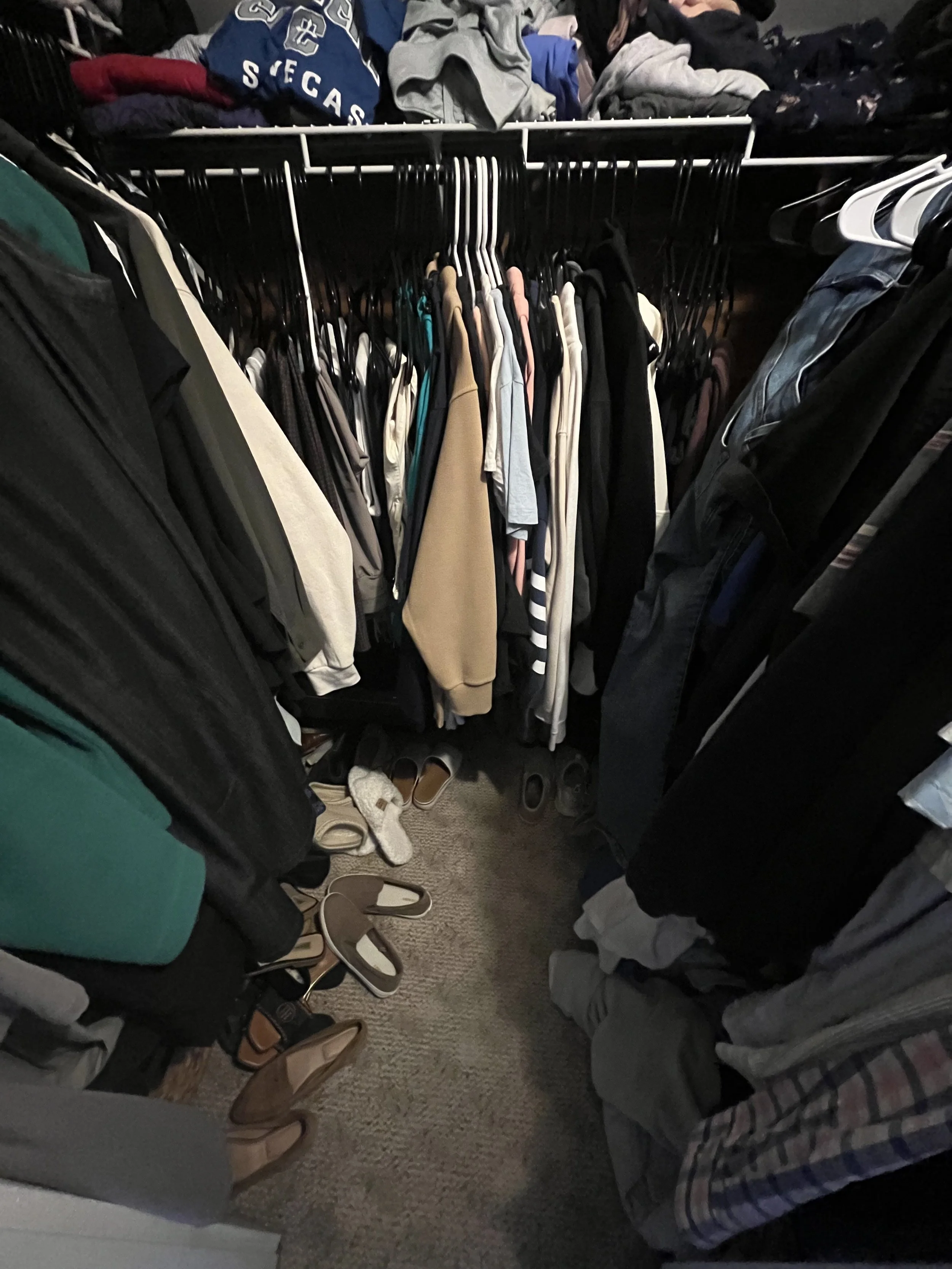 Clothing closet with hanging shirts, pants, and jackets, and assorted shoes and slippers on the floor.