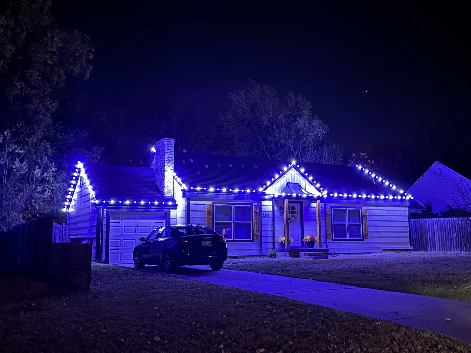 A house decorated with blue Christmas lights at night, with a black car parked in the driveway, two flower pots near the front door, and a chimney visible on the roof.