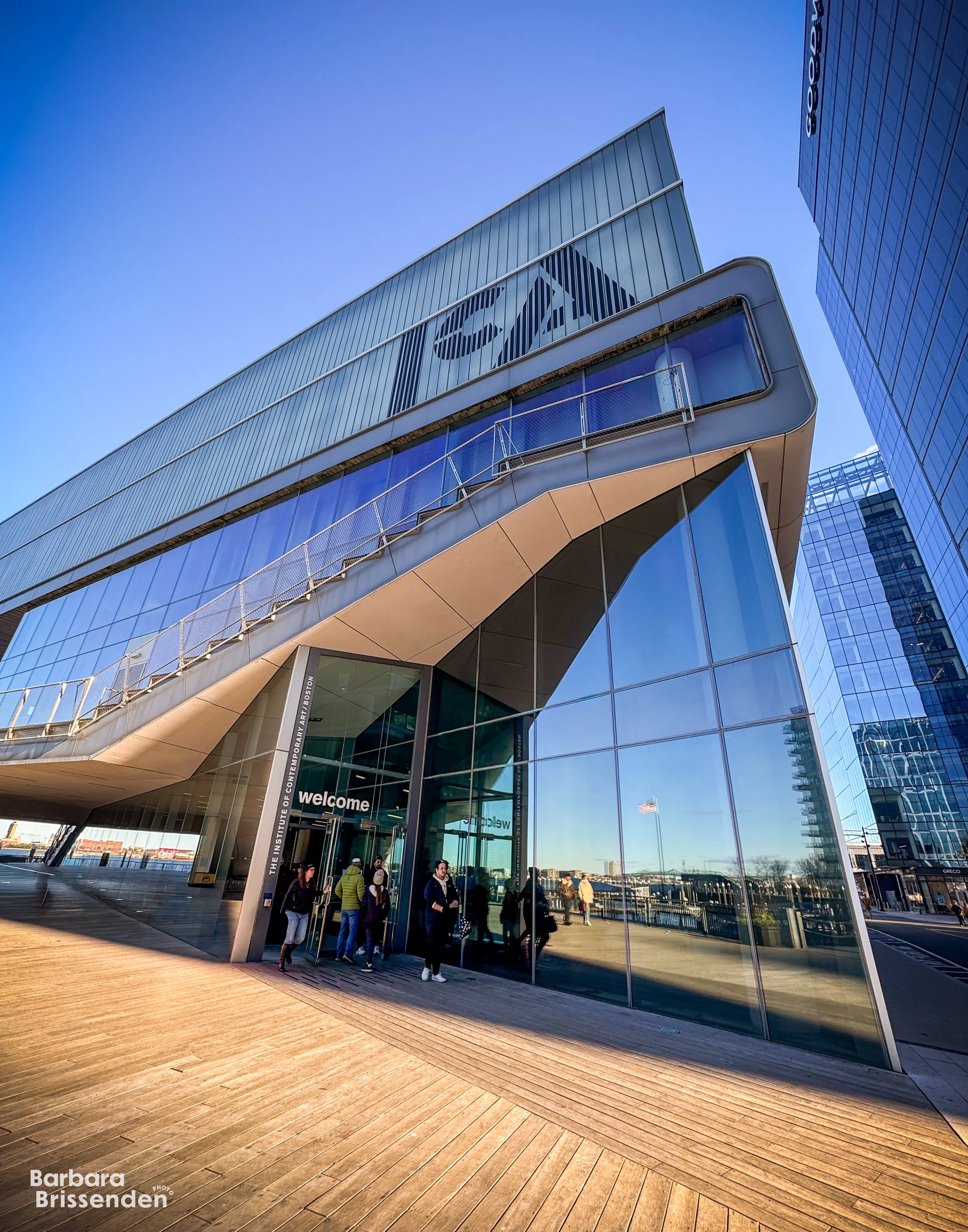 Institute of Contemporary Art Museum with people entering the entrance labeled 'welcome', reflections on the glass, and the ICA logo displayed on the upper part of the building.