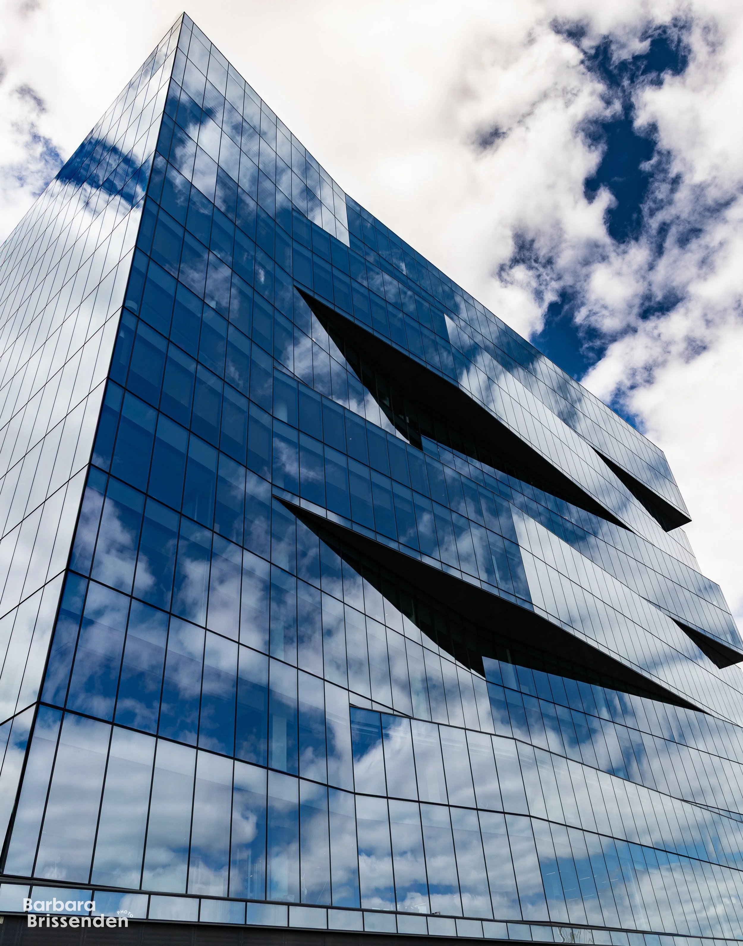 A modern glass office building in the Boston Seaport reflecting the sky with clouds and featuring unique black triangular window accents.