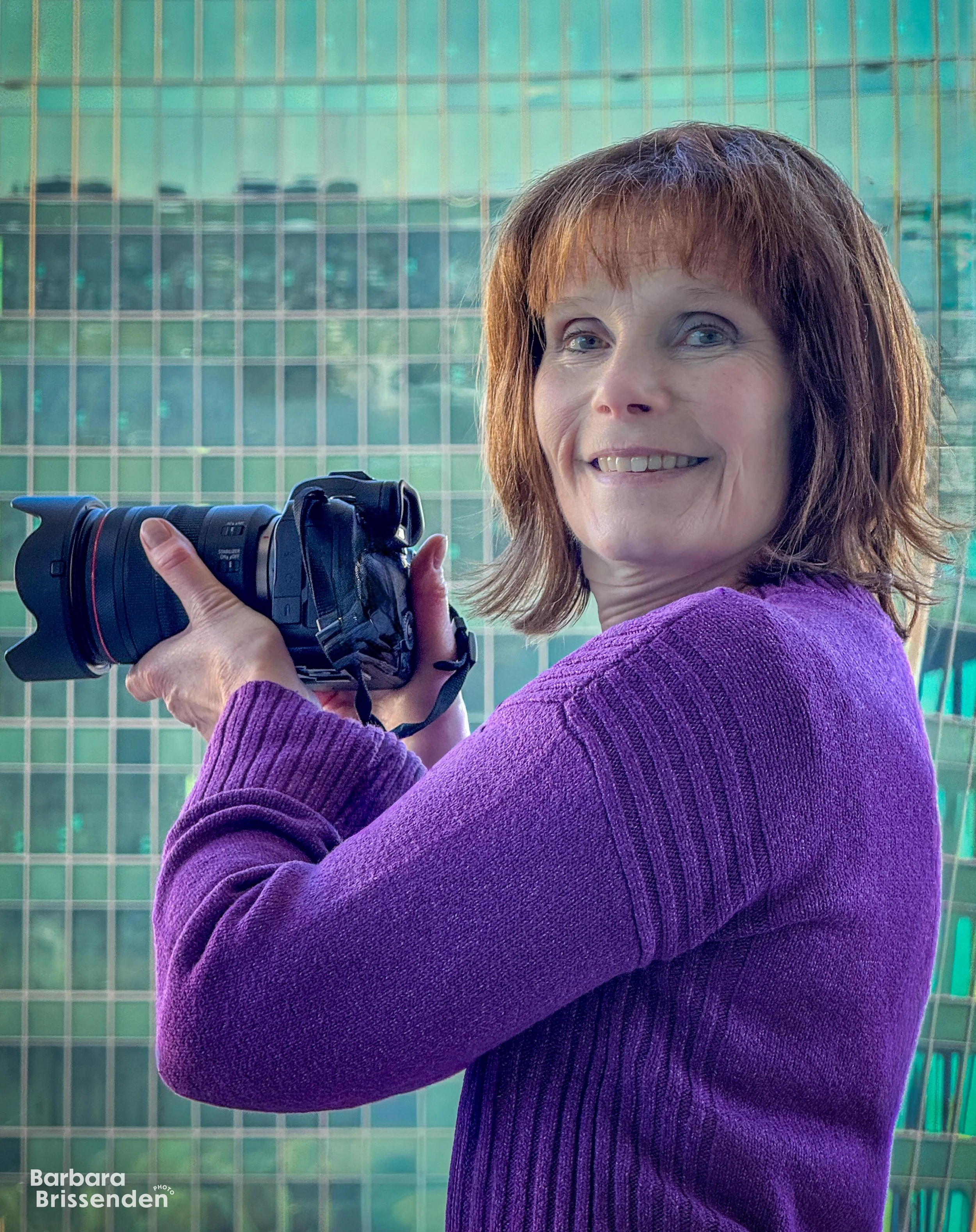 Barbara Brissenden with red hair wearing a purple sweater holds a camera outdoors with a glass building behind her.