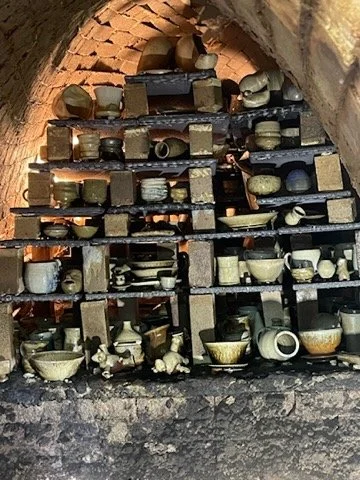 Multiple shelves filled with numerous pottery and ceramic bowls, jars, and vases inside a brick archway. This kiln was built by Ben Owen III.
