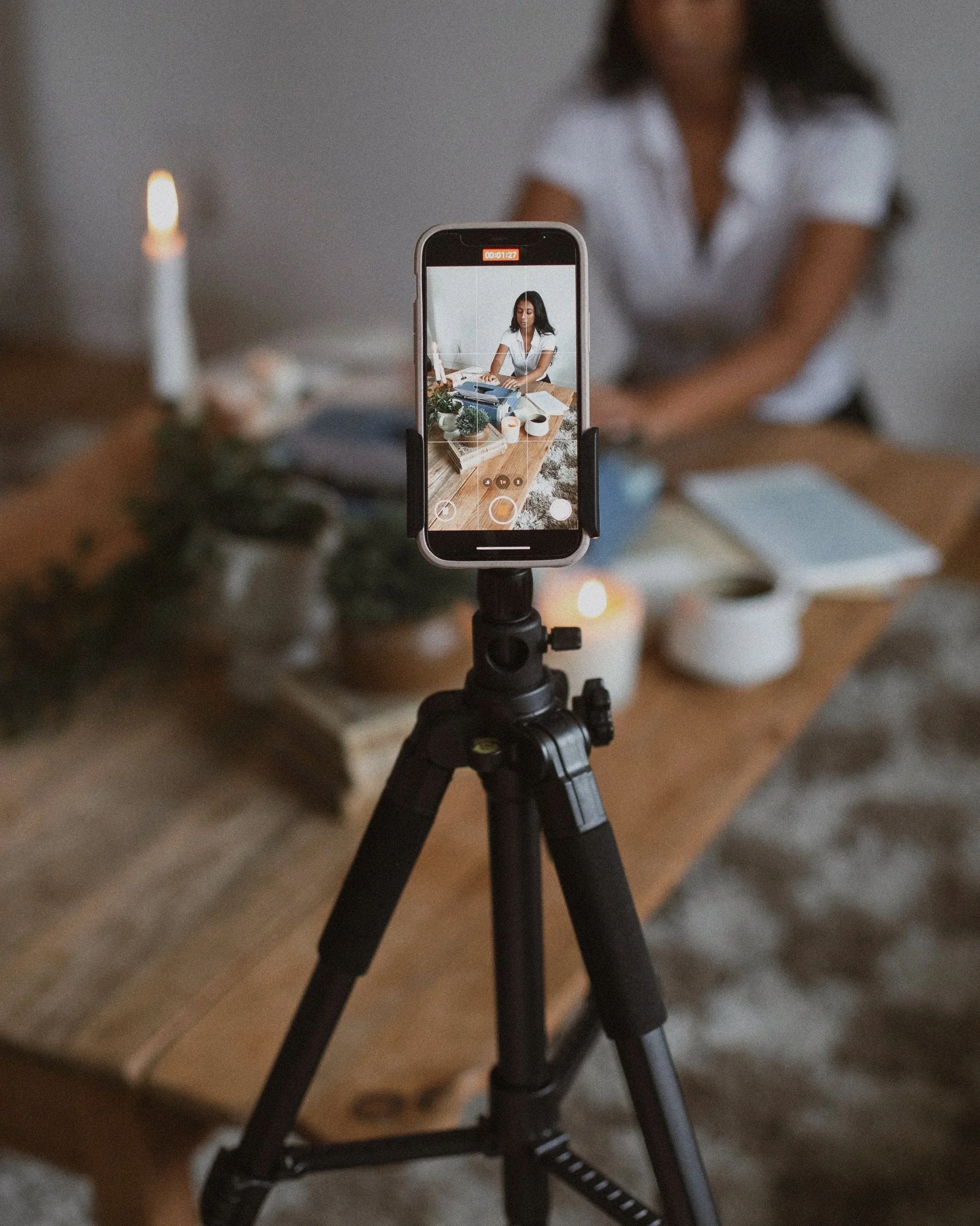 A woman is being filmed at a wooden table with candles, notebooks, and cups, using a smartphone mounted on a tripod.
