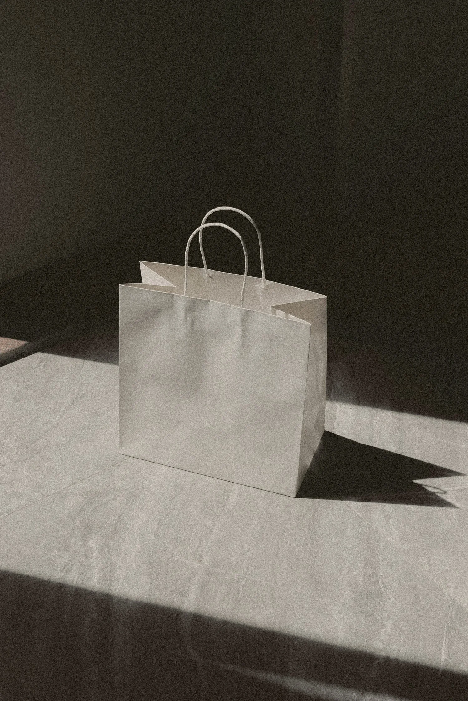 A white shopping bag with handles sitting on a tiled floor, shadows cast by lighting on the bag and floor.