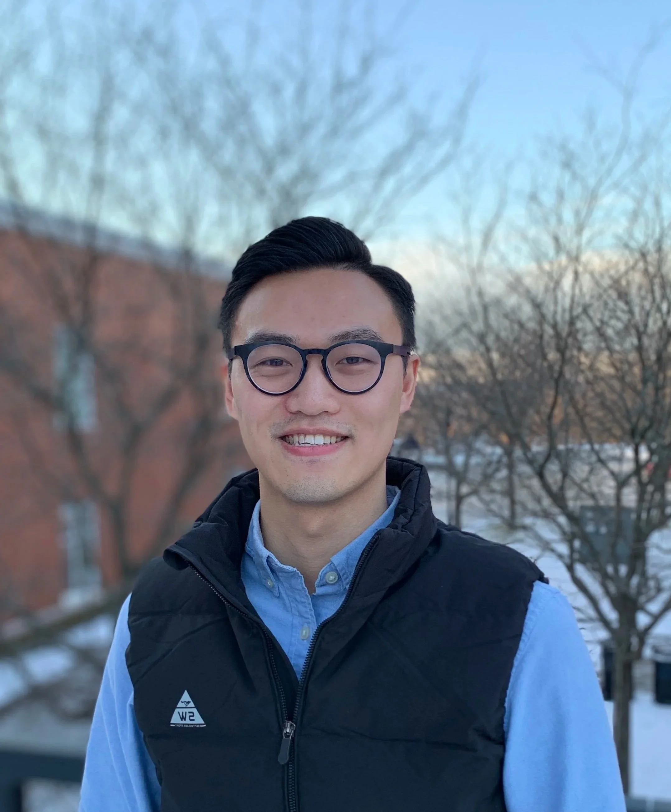 A young man with glasses and short black hair smiling outdoors against a background of leafless trees and a building during winter.