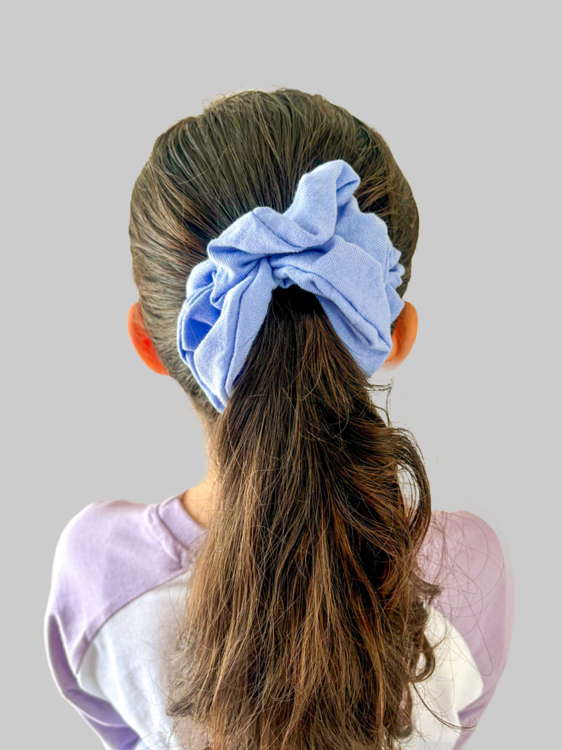 Back view of a girl with long brown hair tied with a large, light blue scrunchie, wearing a light purple shirt, against a plain light gray background.