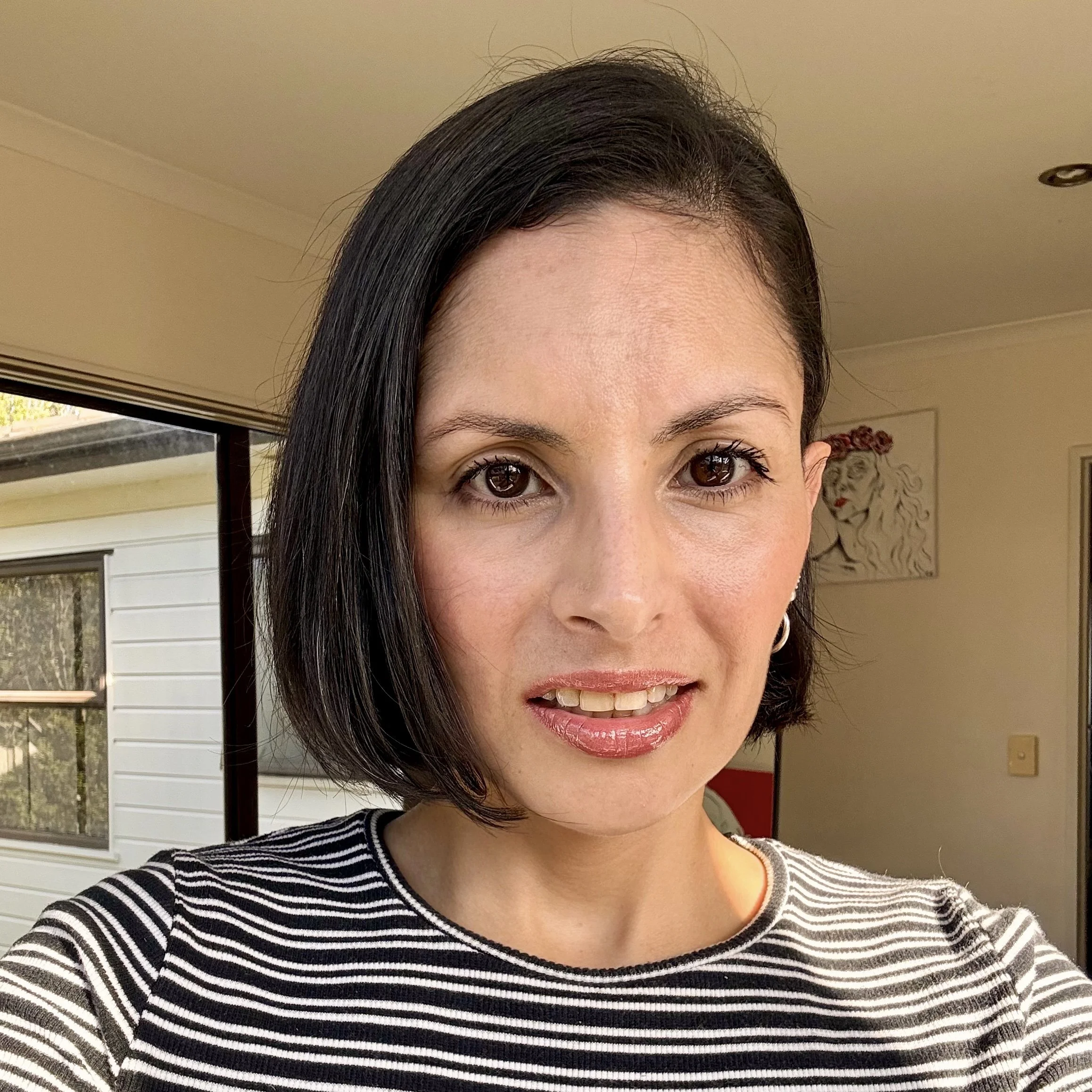 Close-up of a woman with straight, dark hair and brown eyes, wearing a black and white striped shirt, standing indoors near a window.