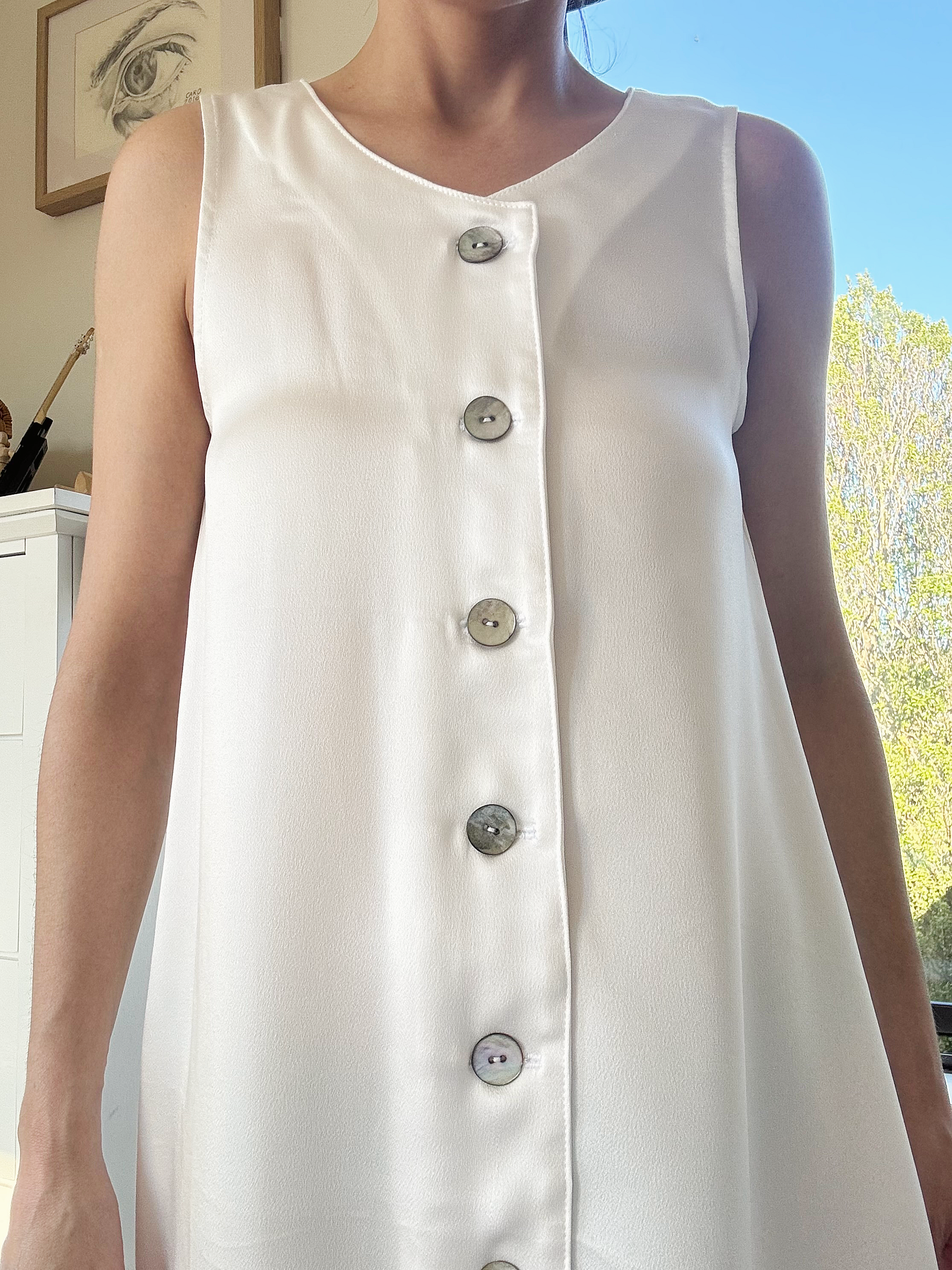 A woman wearing a sleeveless white dress with large mother of pearl buttons down the front, standing indoors near a window with sunlight and a view of trees outside.