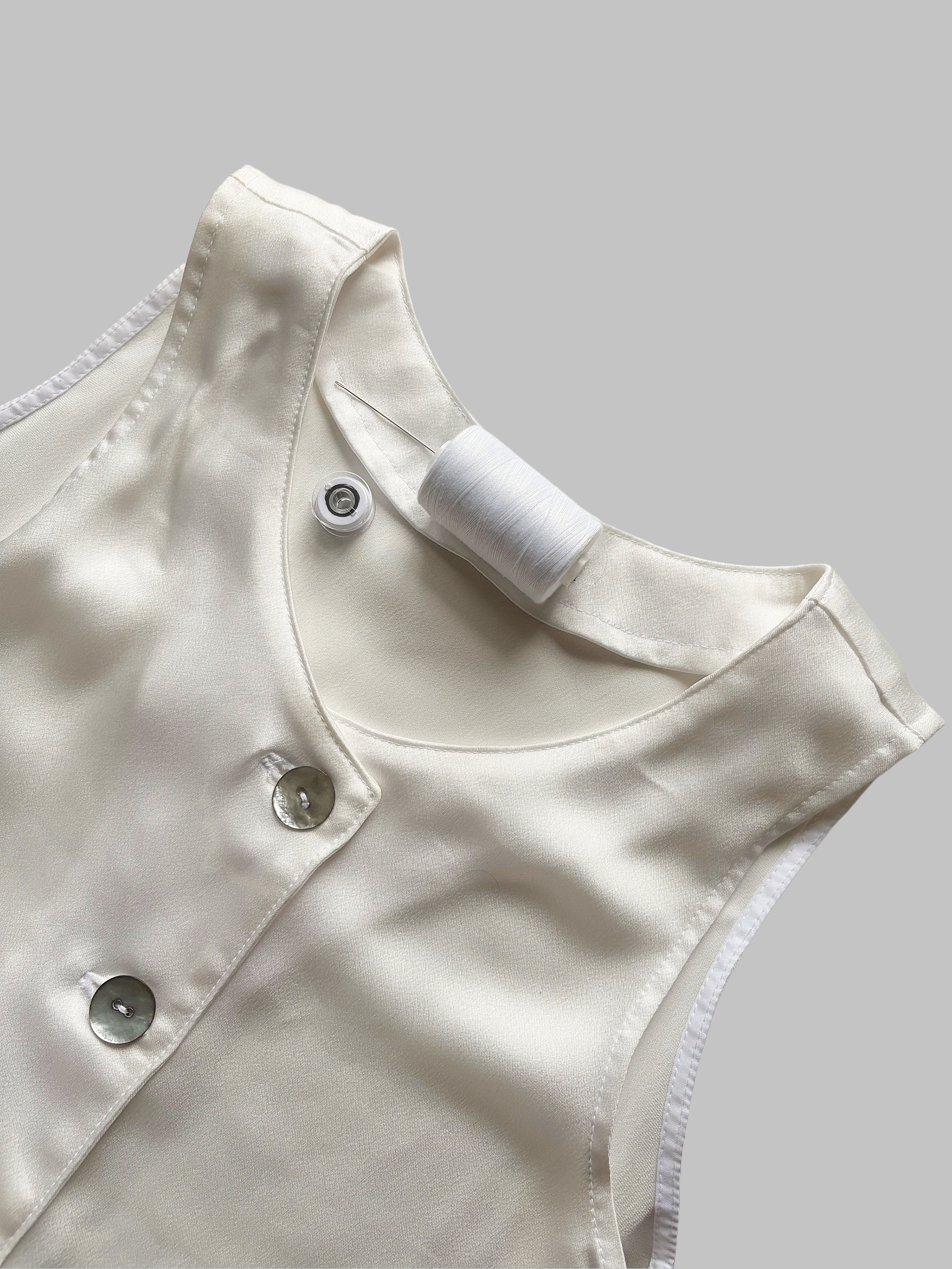 Close-up of an ivory satin sleeveless dress with shell buttons sewn, a stitching thread, a machine bobbin and a hand sewn needle placed on top.
