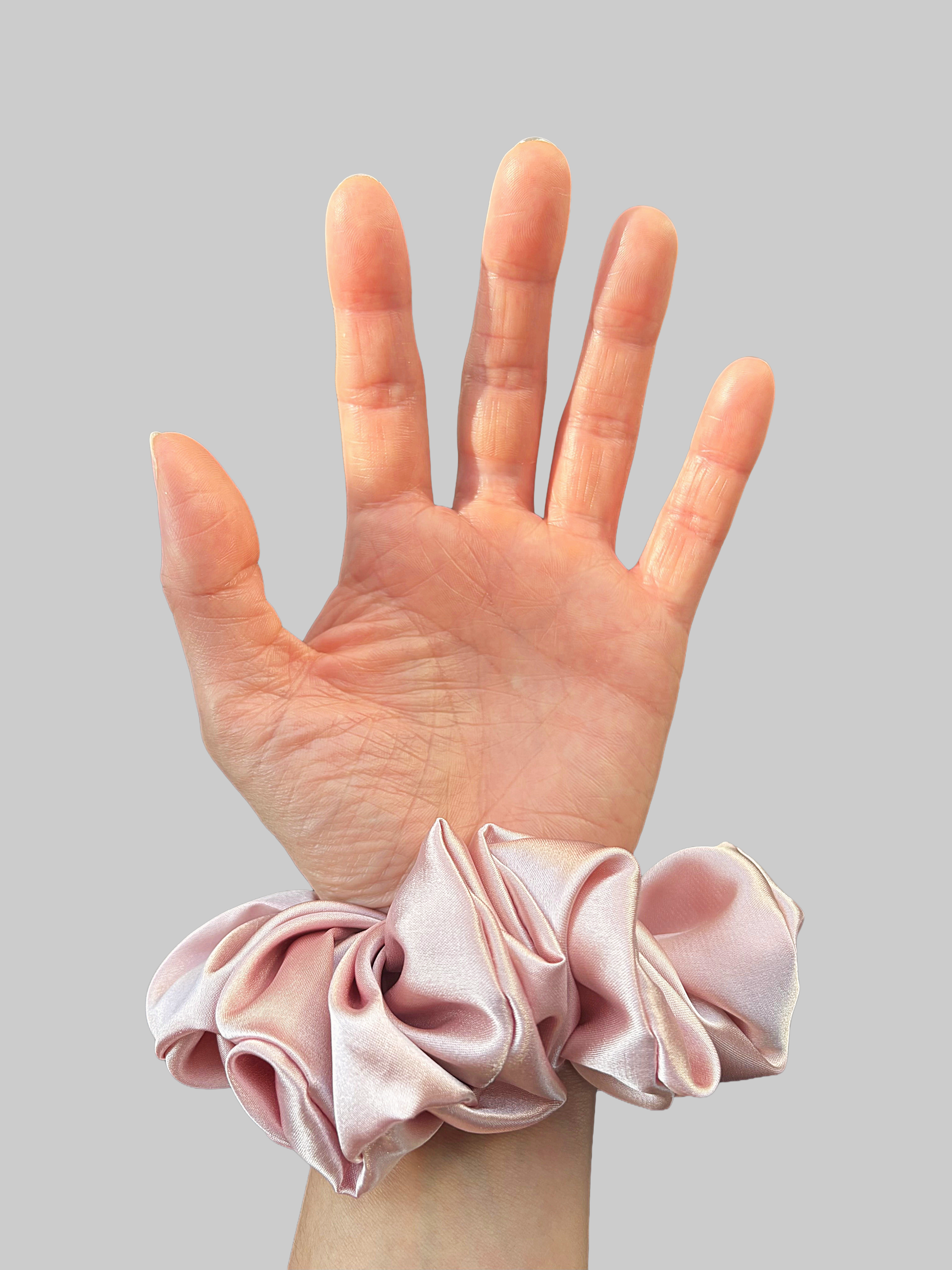 Close-up detail of a woman's hand with fair skin tone against a grey background. The wrist is adorned with a pink satin scrunchie.