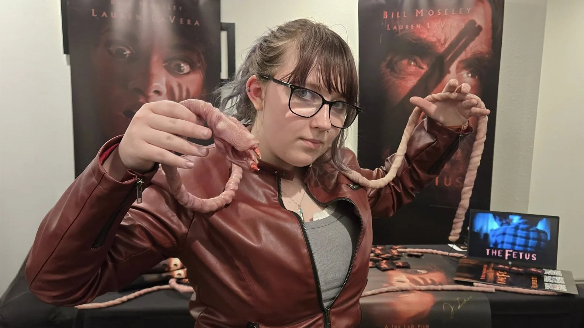 Horror fan posing with The Fetus creature prosthetic at an indie horror film booth during a horror convention.
